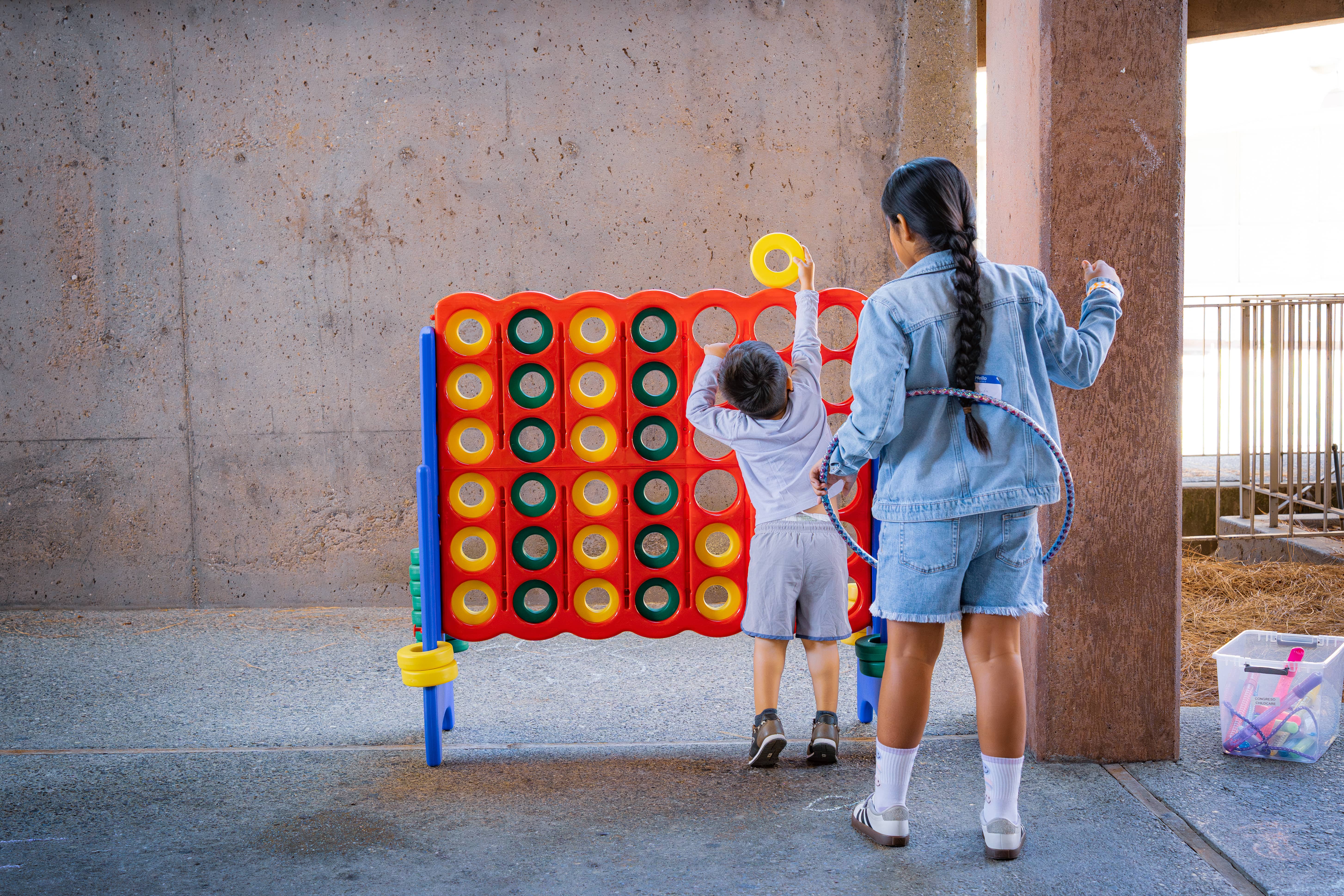 Two children playing a large Connect Four game, one child reaching to place a yellow disc while the other holds a hula hoop around their waist.
