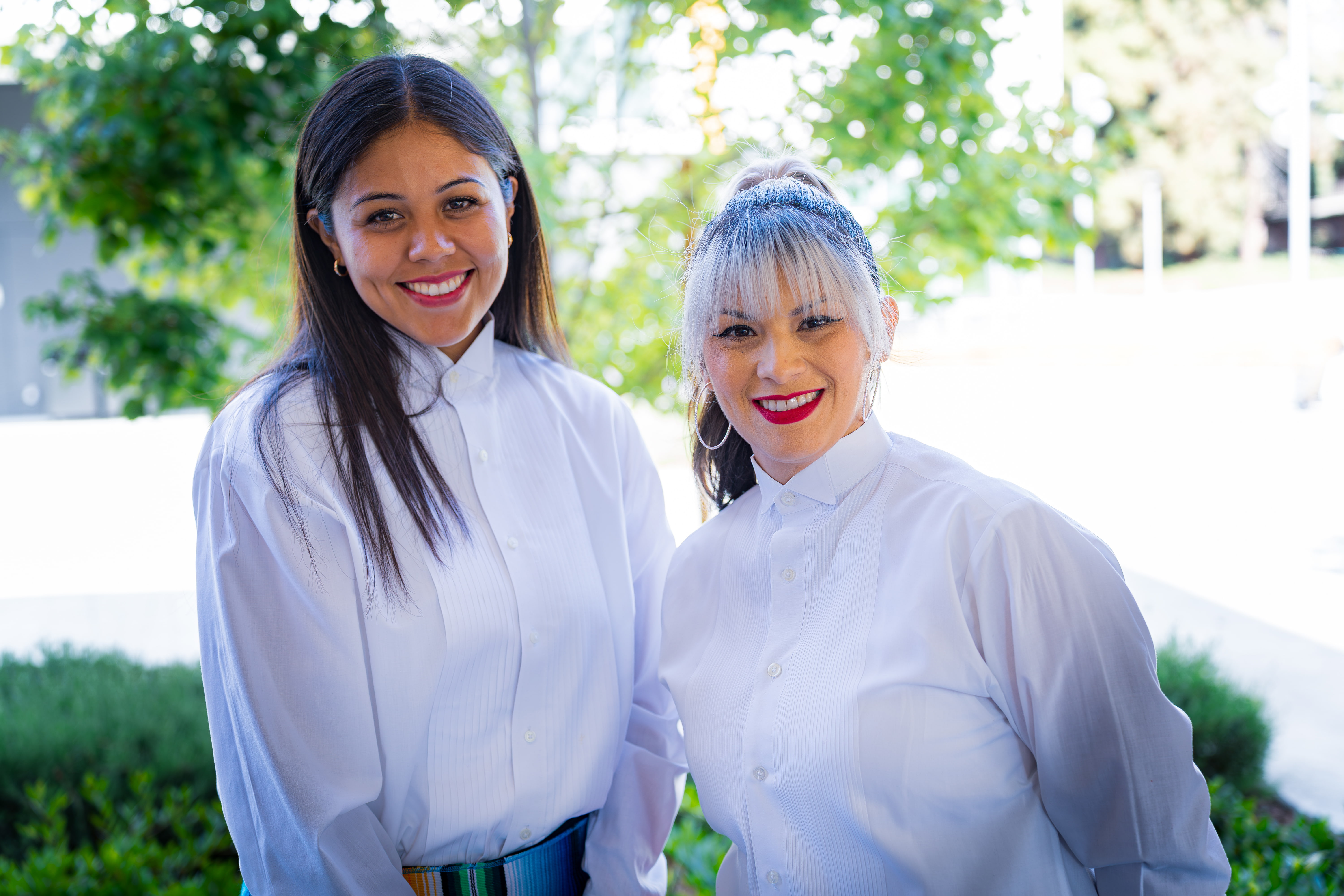 Two women smiling outdoors wearing white button-up shirts with greenery in the background.