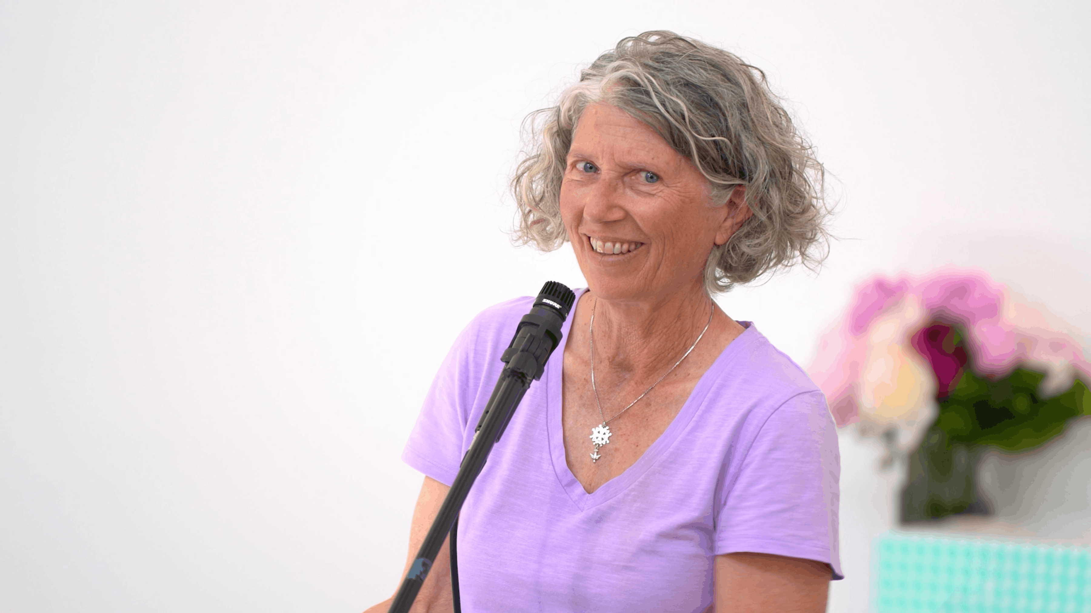 Smiling older woman with curly gray hair wearing a lavender shirt, speaking into a microphone with blurred flowers in the background.
