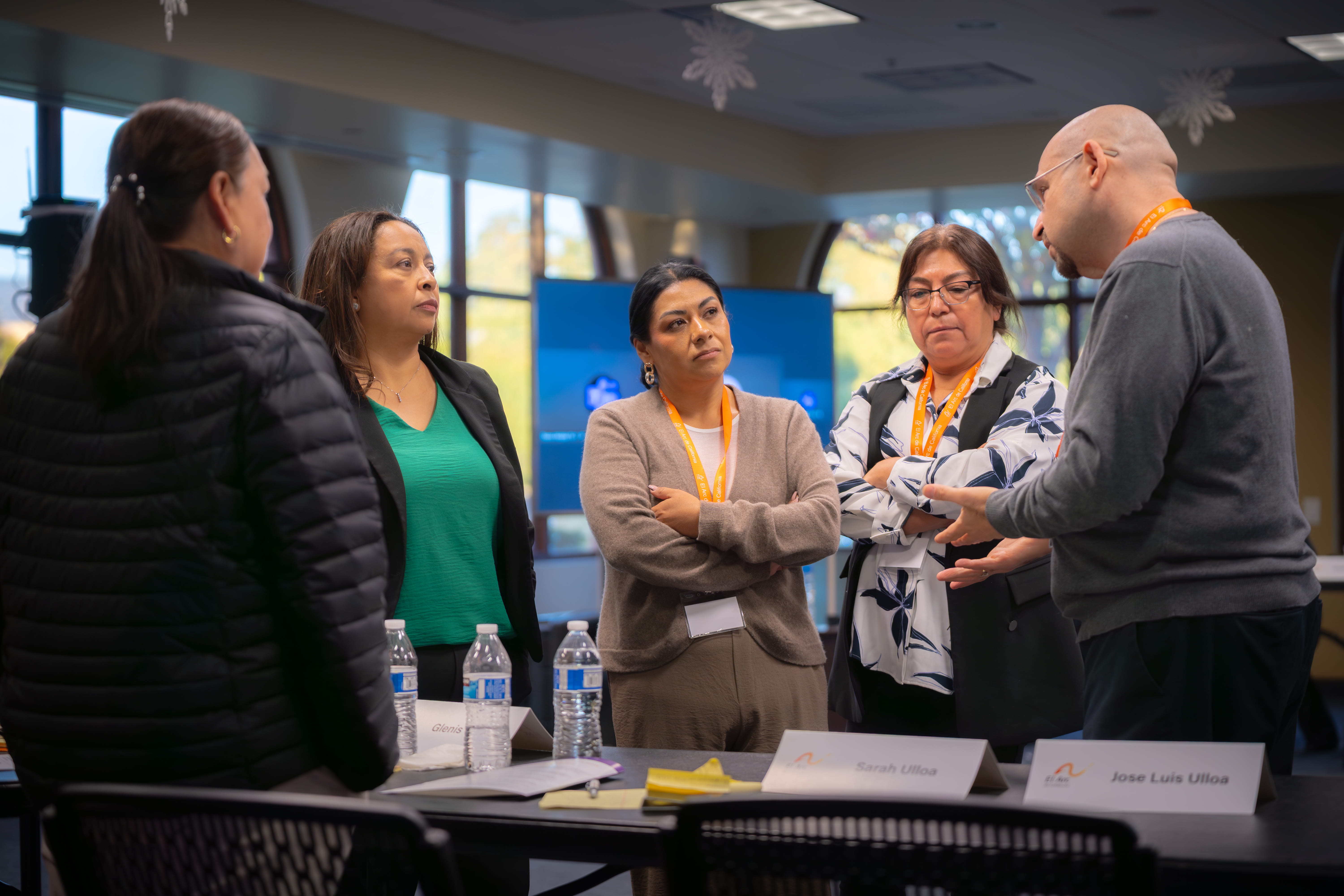 Five adults engaged in a serious discussion around a conference table with name tags and water bottles visible.