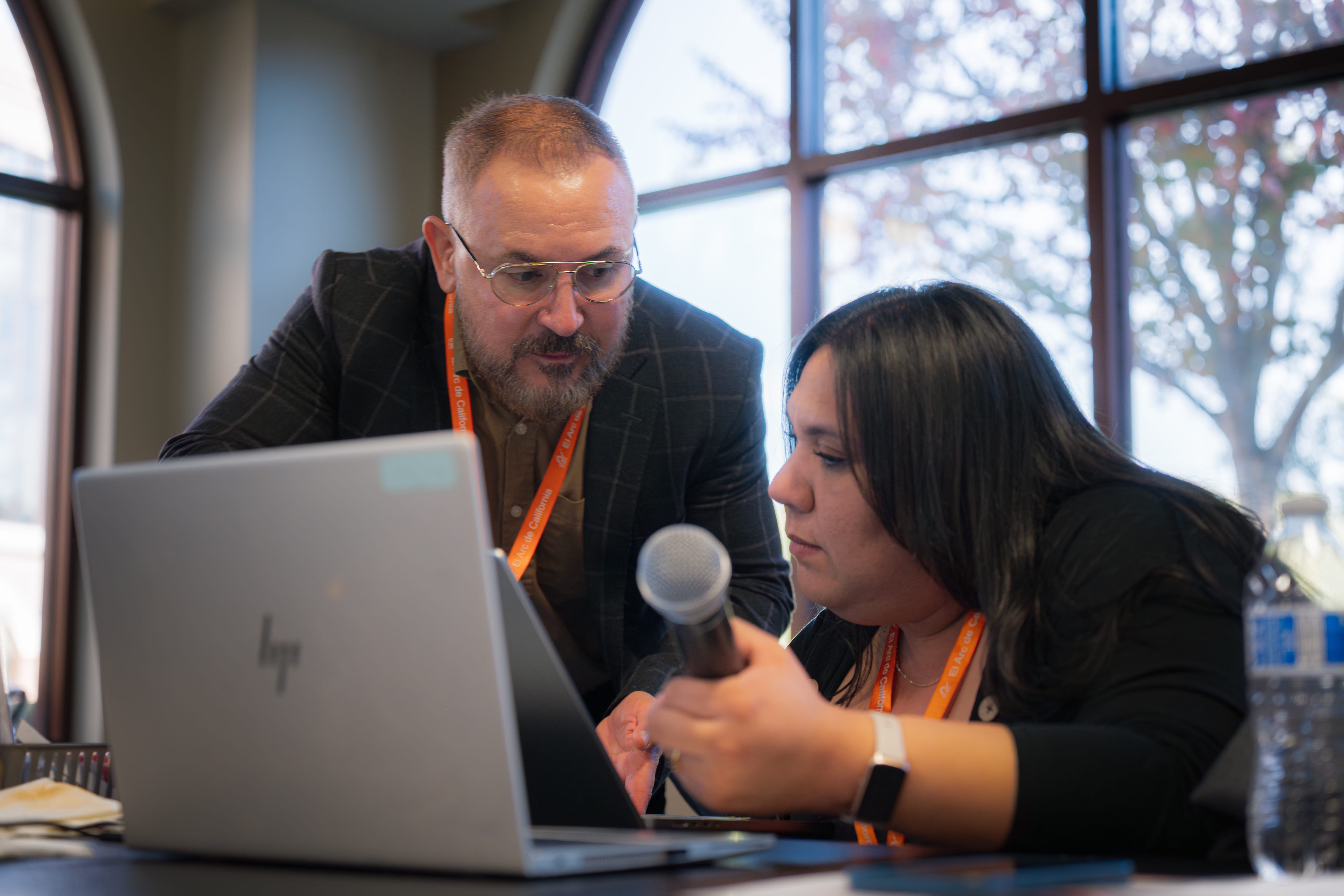 Two people working together on a laptop in a room with large windows, one holding a microphone.