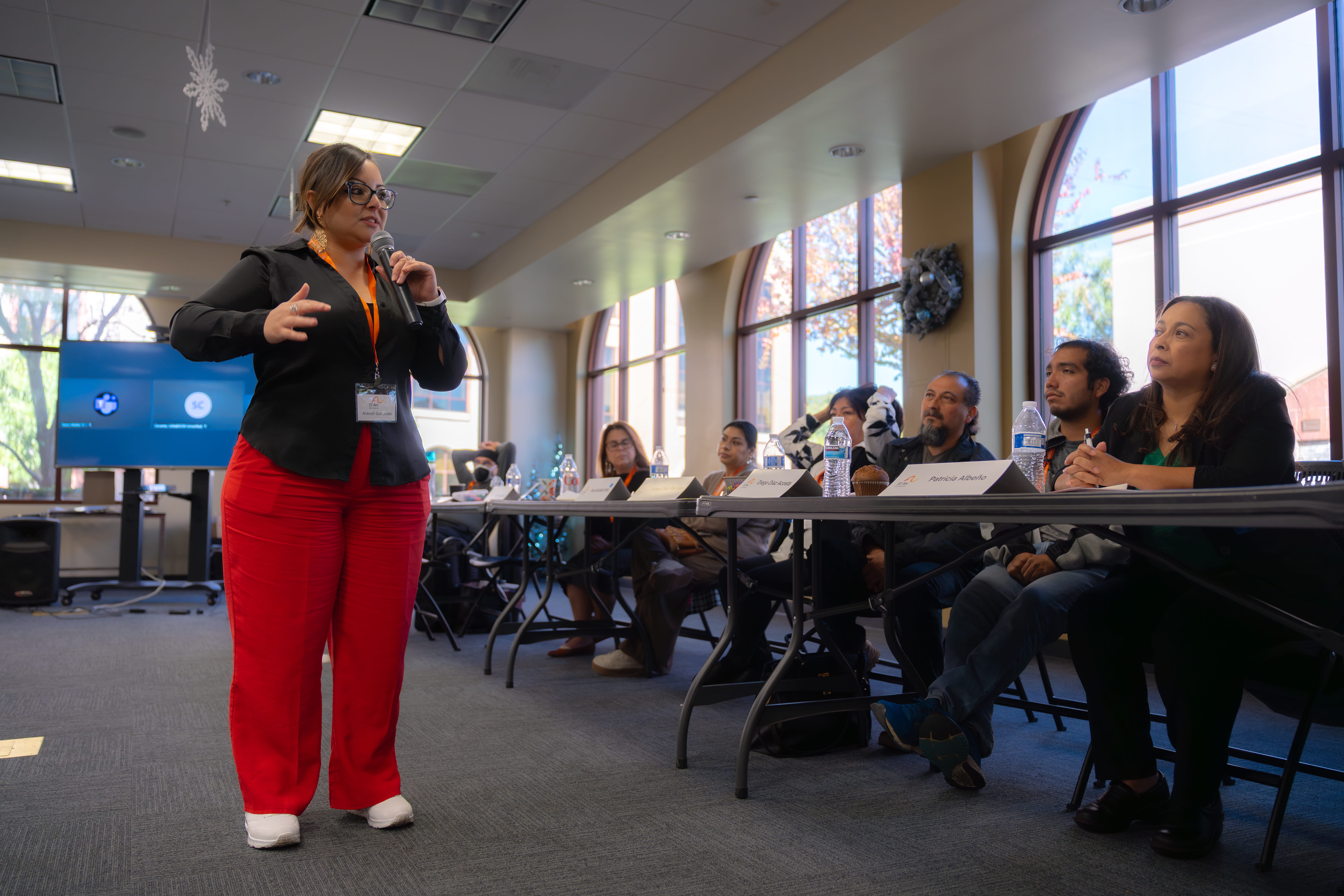 Woman with glasses and red pants speaking into a microphone to an attentive panel seated at a long table in a conference room with large windows.