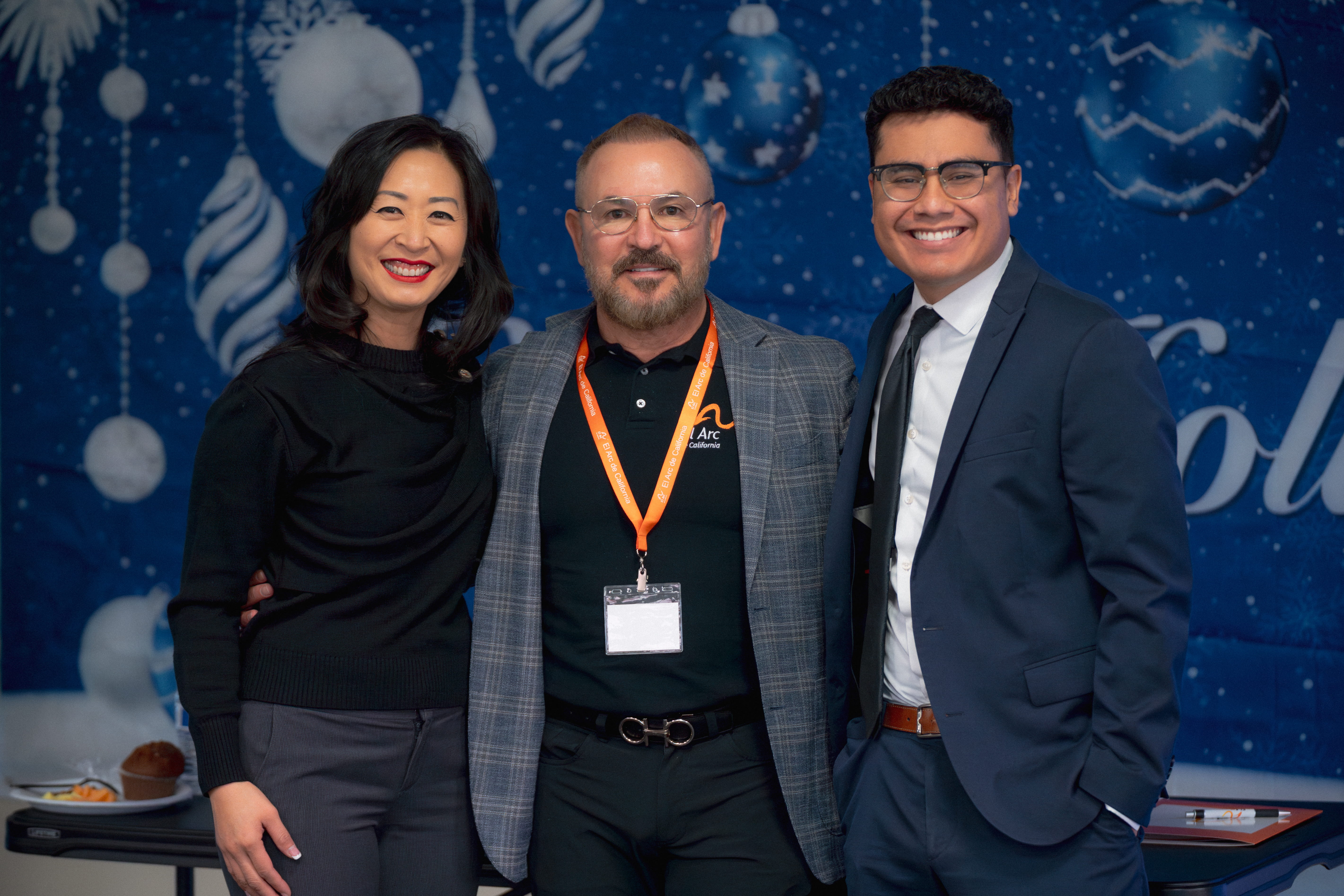 Three people smiling and posing together in front of a blue holiday-themed backdrop with white ornaments.