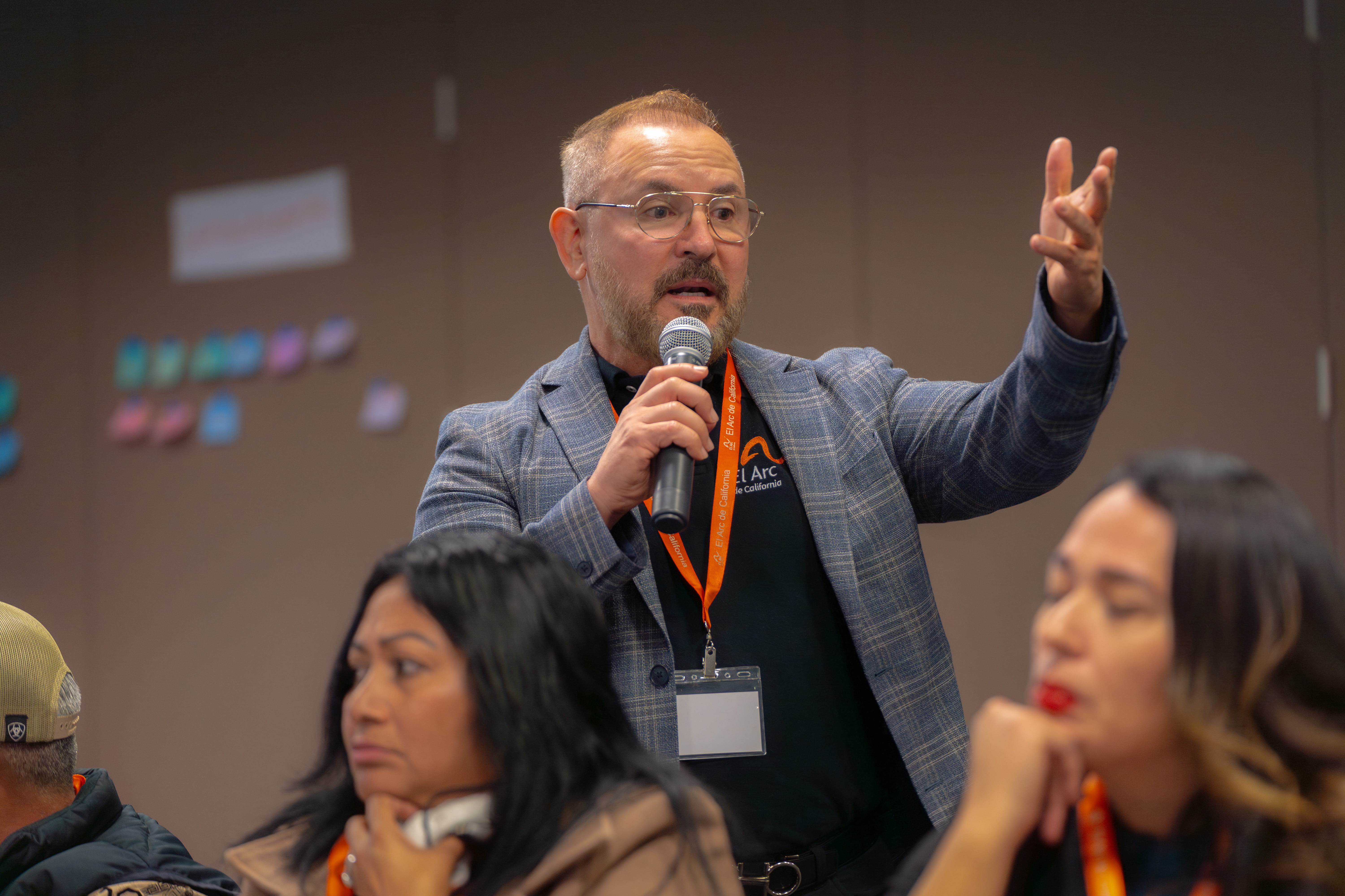Man with glasses and a beard speaking into a microphone and gesturing with his right hand during a meeting.