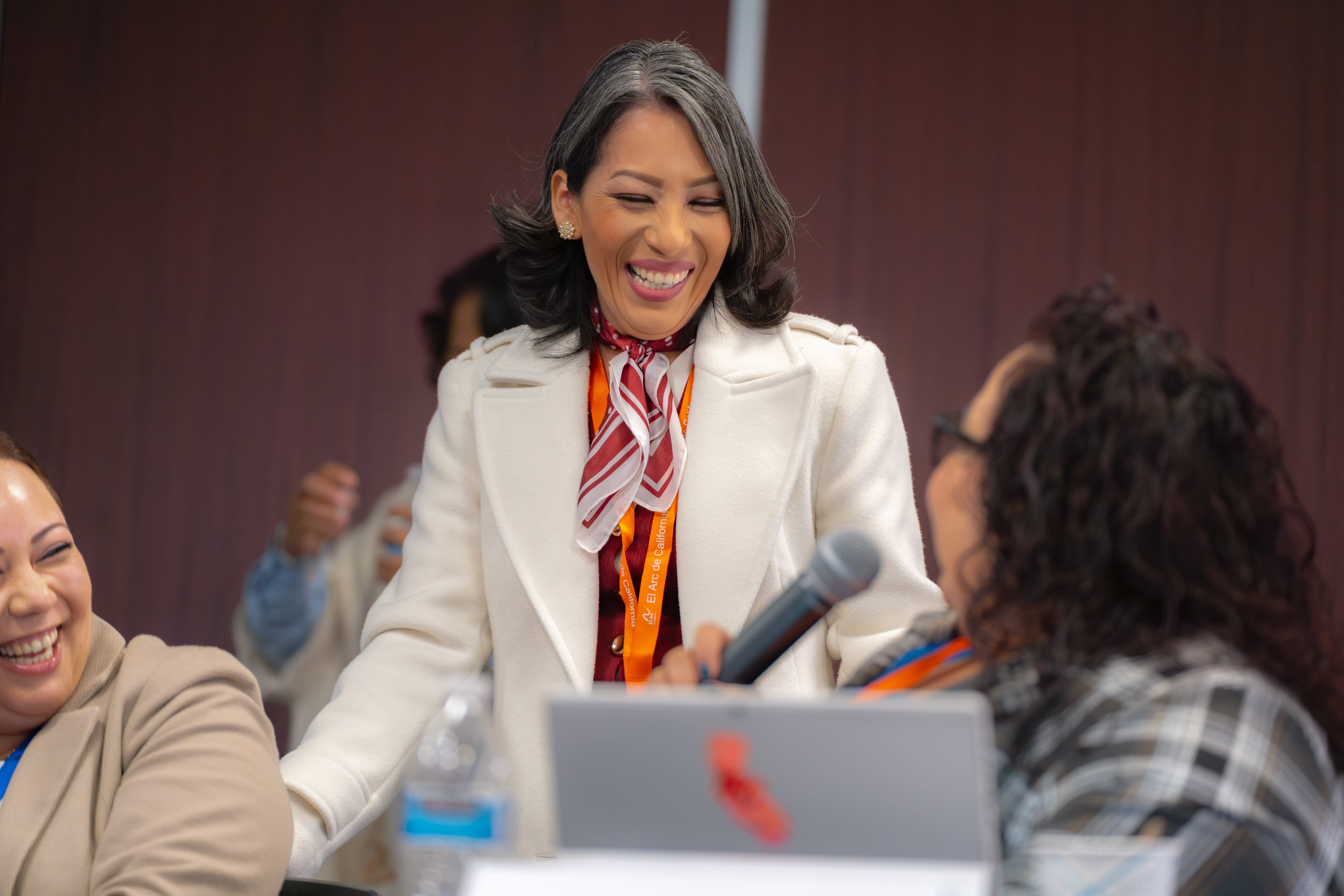 Smiling woman in white coat and striped scarf interacting with seated women, one holding a microphone.