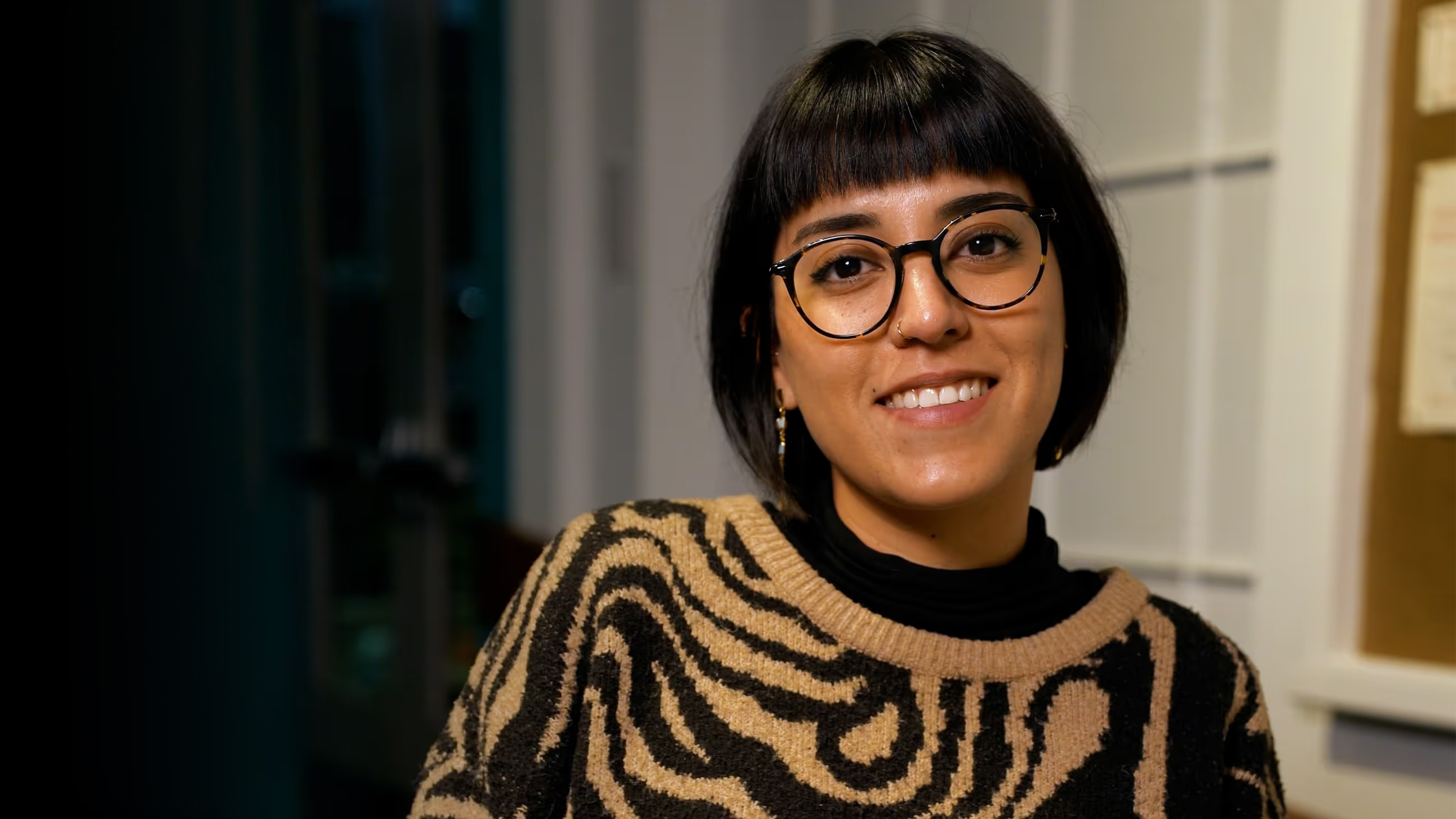 Latina woman looking at camera with glasses and a bob haircut with a cool sweater (brown and black swirls in a pattern).

