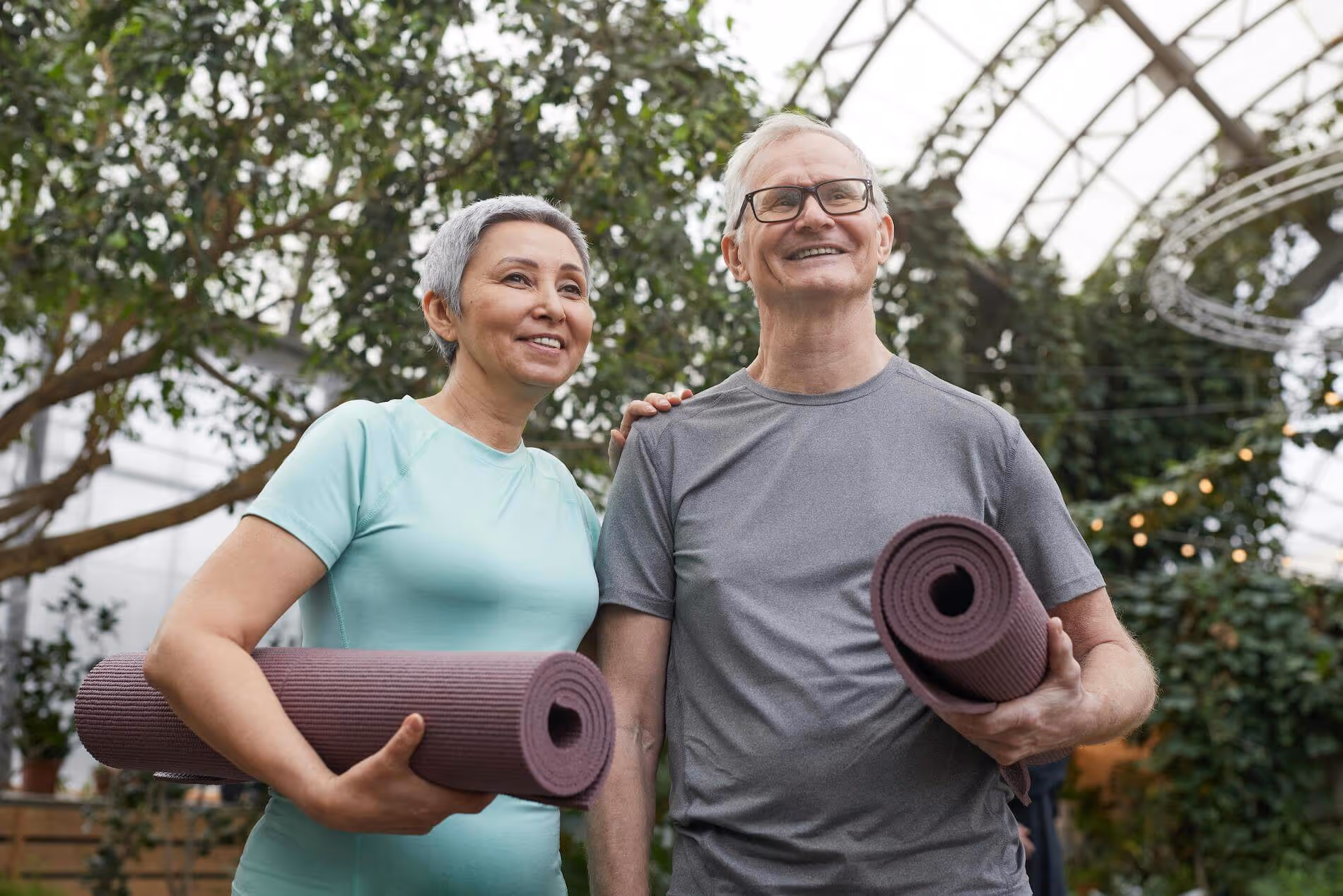 Smiling elderly man and woman holding rolled yoga mats in a bright indoor greenhouse with plants.