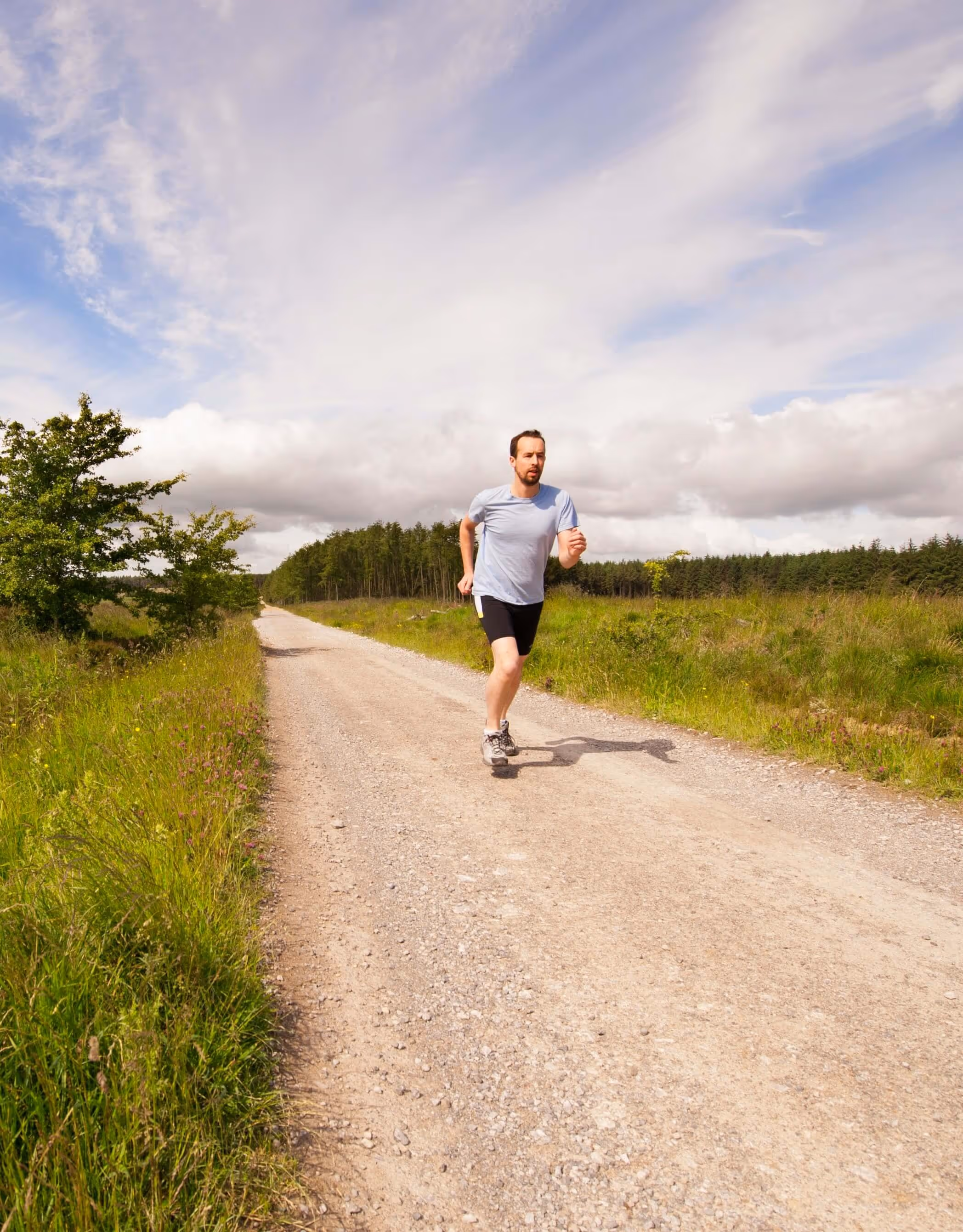 Man jogging on a gravel path surrounded by green grass and trees under a partly cloudy sky.