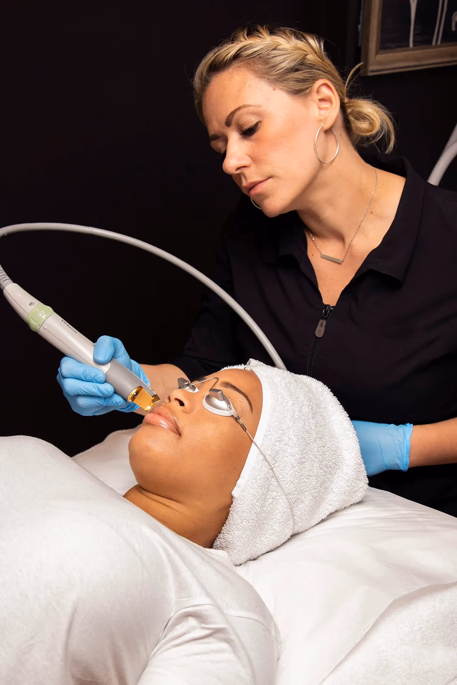 Esthetician wearing blue gloves performing a facial laser treatment on a woman lying down with protective eye shields and a towel wrapped around her head.