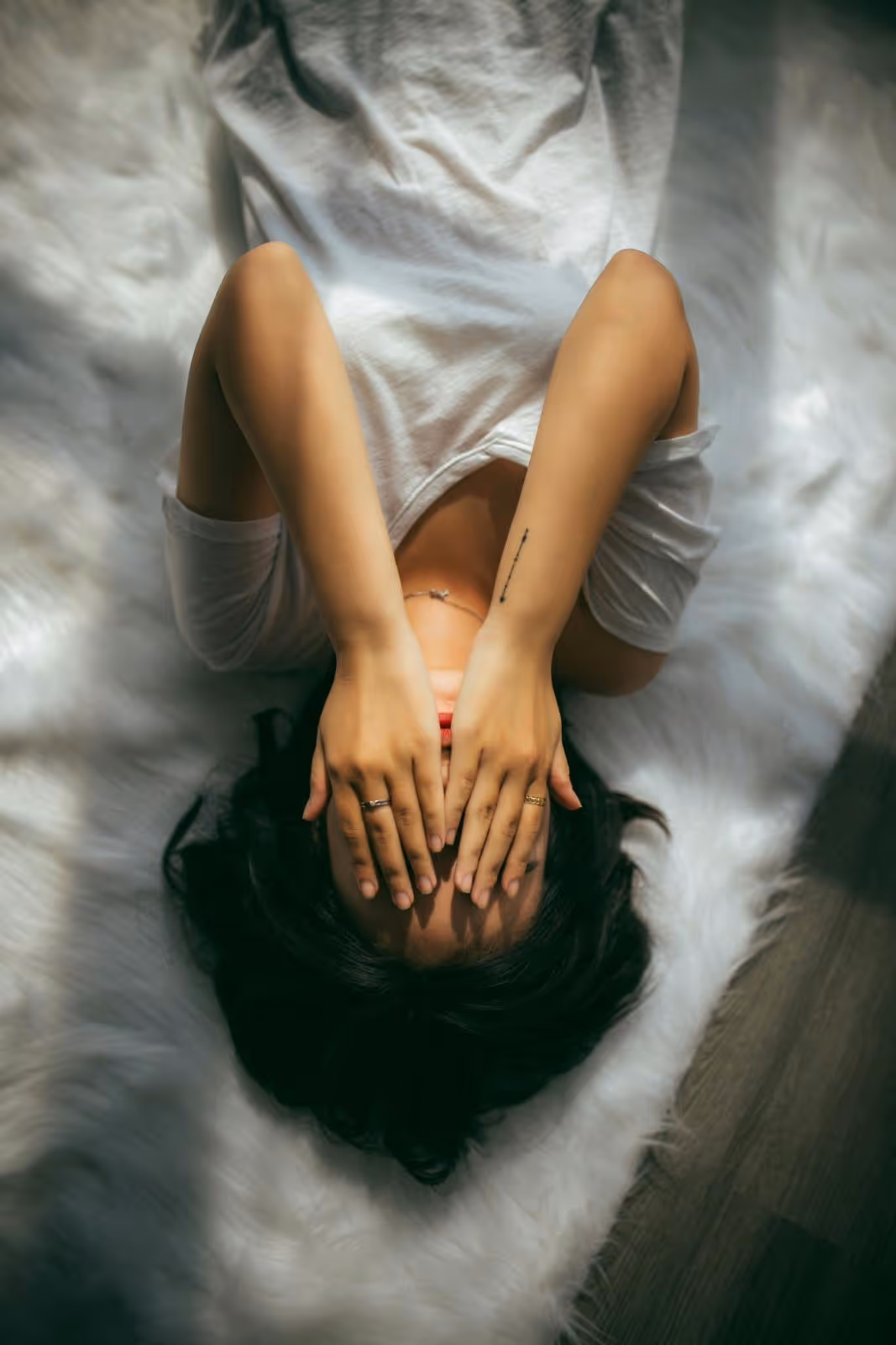 Person lying on white fur rug with hands covering face, wearing a white shirt and rings.