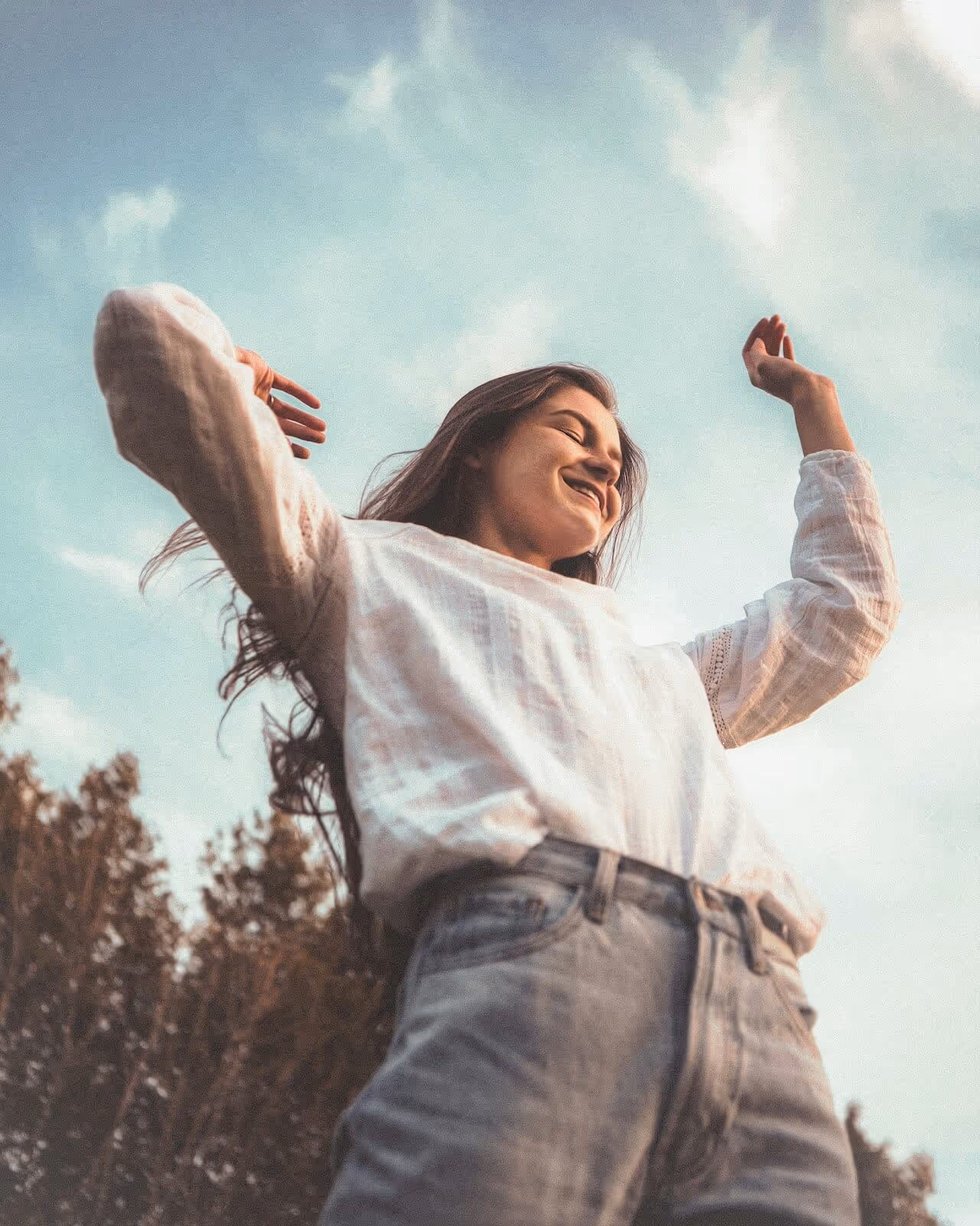 Young woman in a white blouse and jeans smiling with her arms raised outdoors under a blue sky.