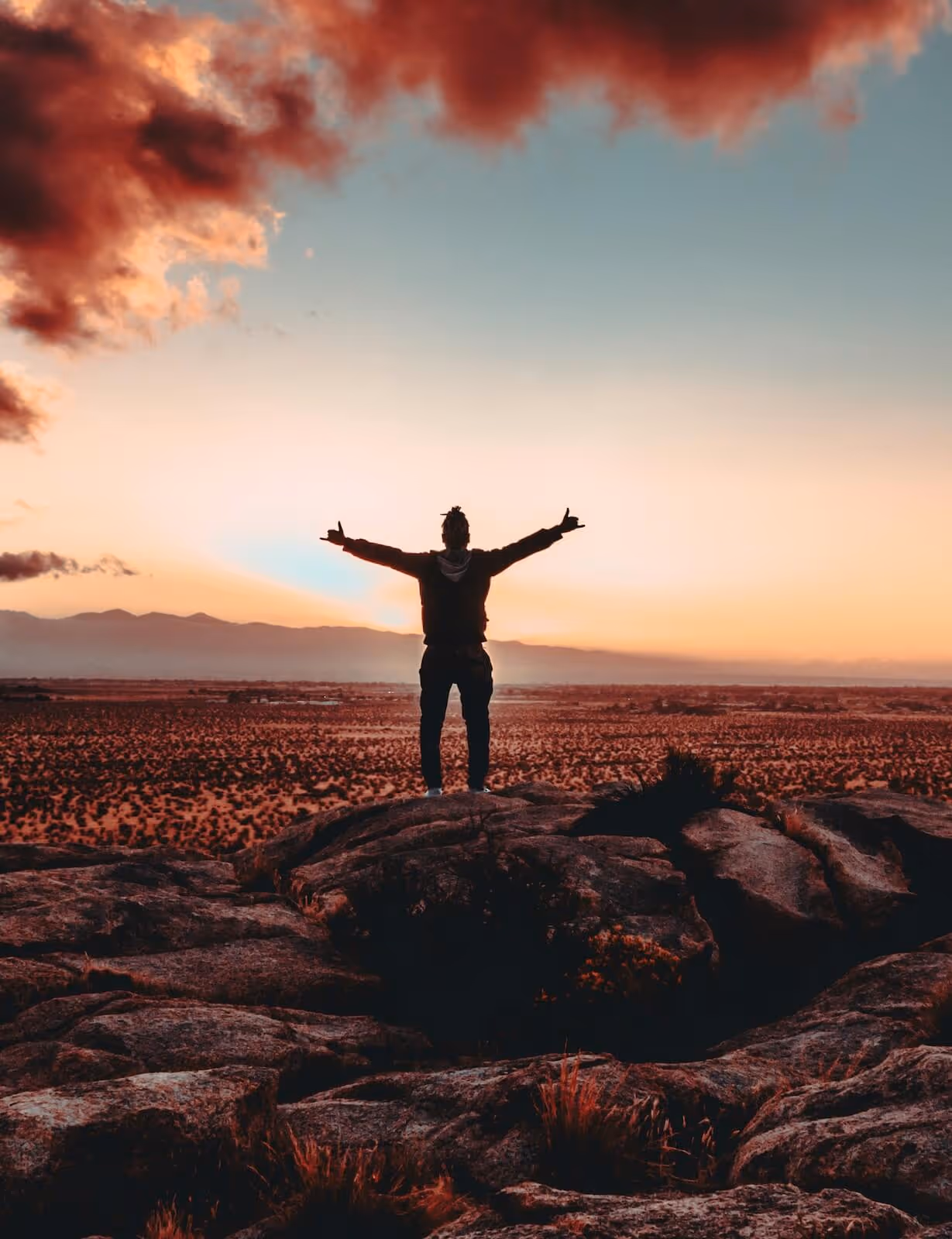 Silhouette of a person standing on rocky terrain with arms outstretched at sunset under a sky with dramatic red clouds.