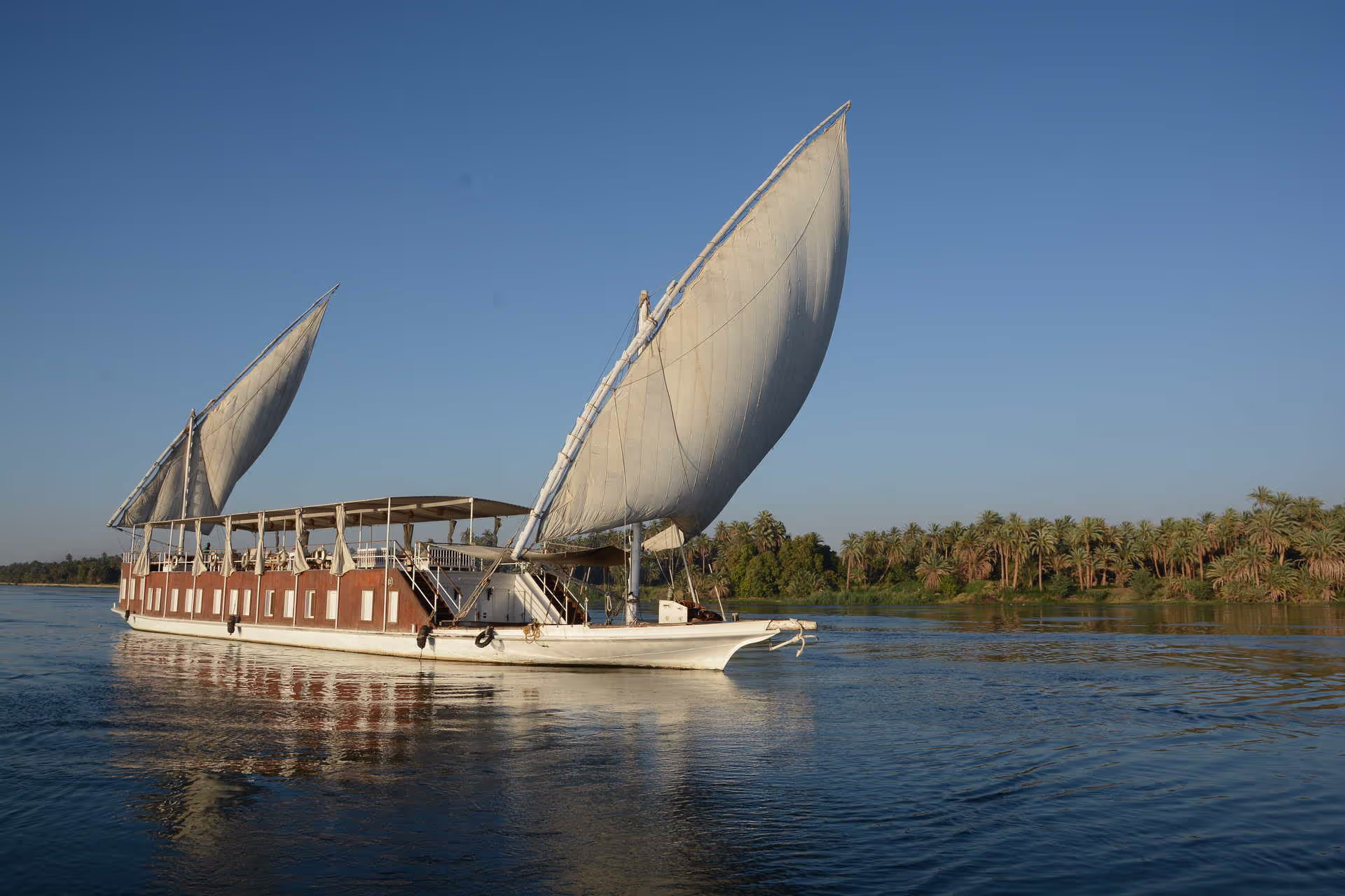 Grand bateau à voile traditionnel naviguant sur une rivière calme avec des palmiers en arrière-plan sous un ciel bleu clair.