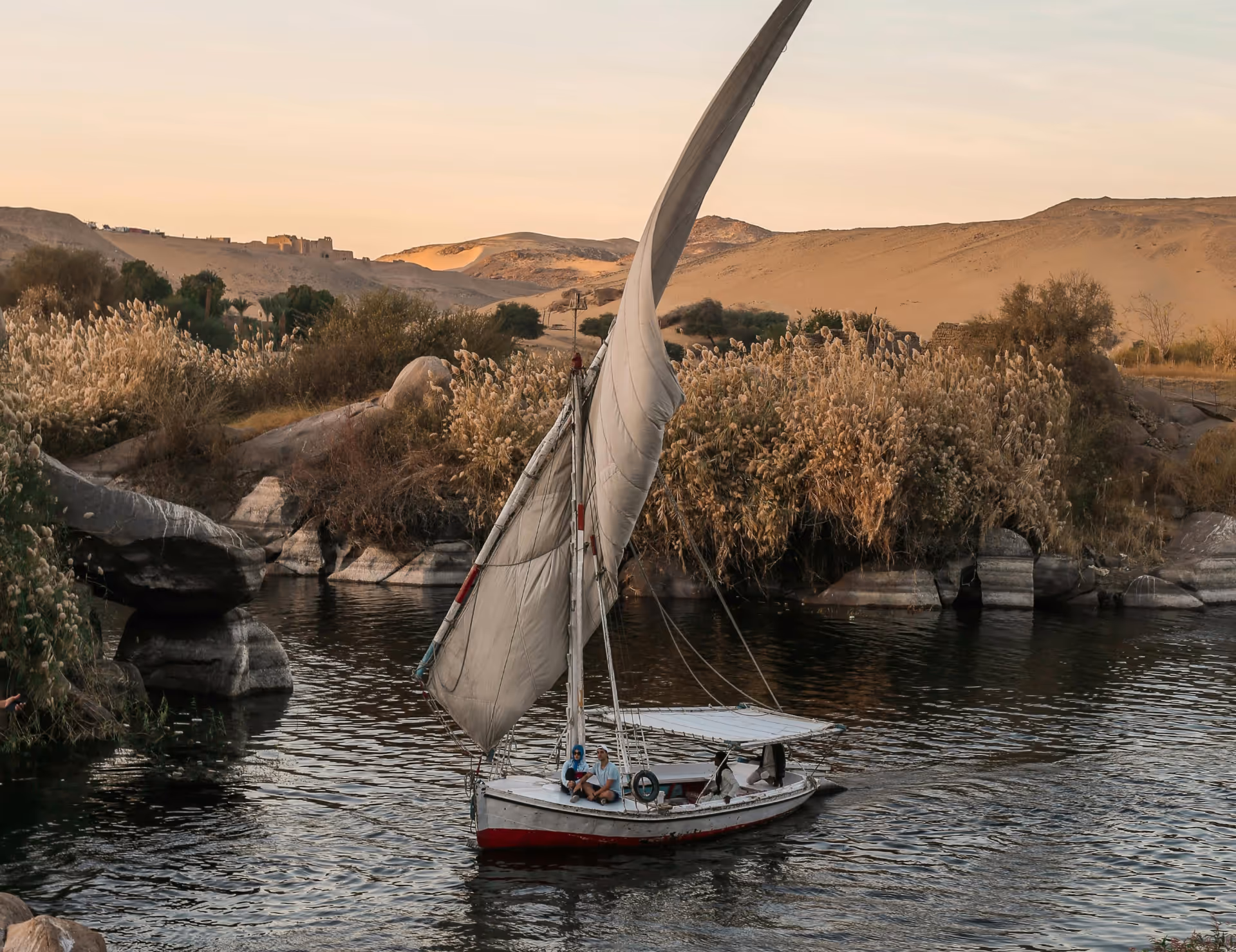Petit bateau à voile naviguant sur une rivière calme entourée de rochers, de roseaux et de collines désertiques au coucher du soleil.