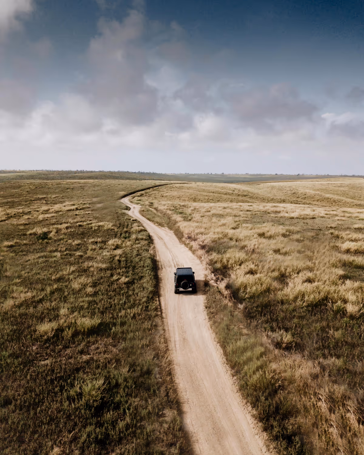 Un véhicule noir circule sur une route de terre sinueuse au milieu de vastes plaines herbeuses sous un ciel nuageux.