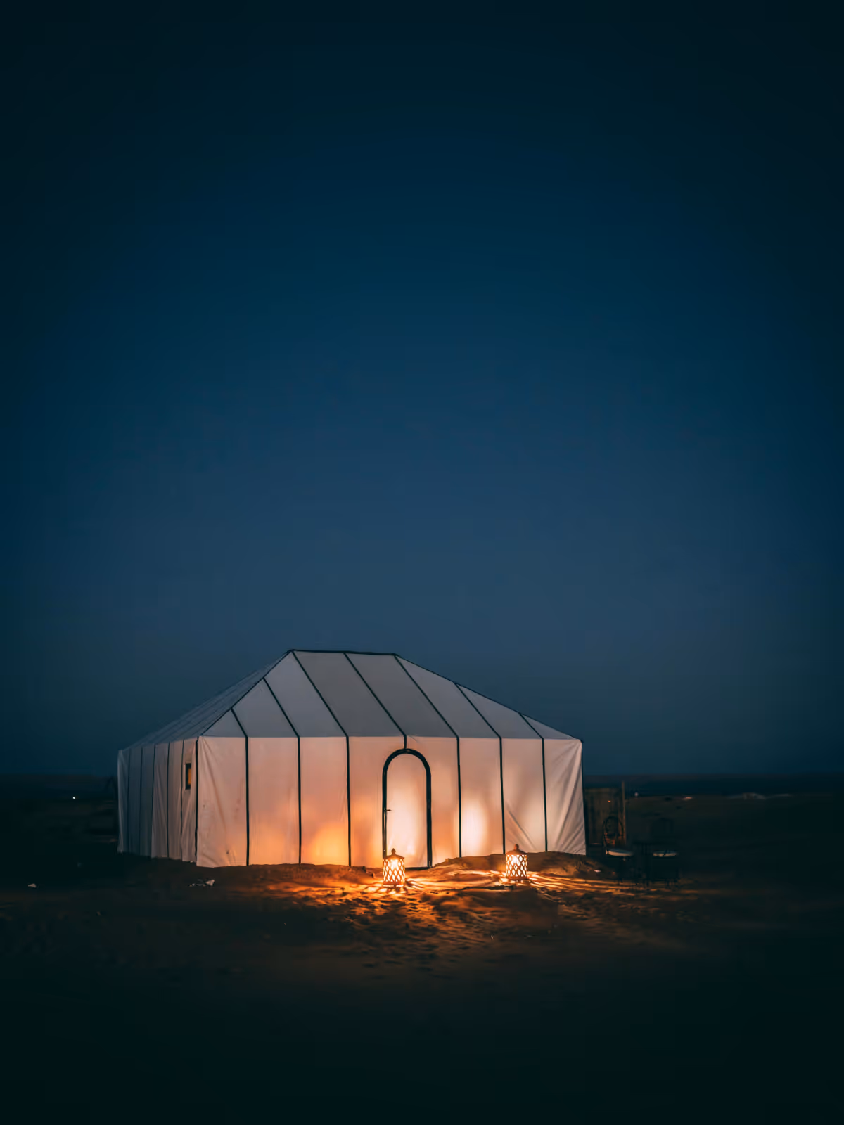 Grande tente blanche éclairée la nuit dans un désert, avec deux lanternes allumées devant l'entrée.