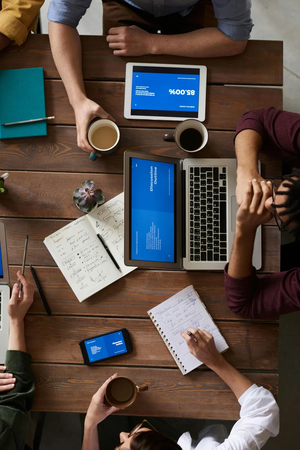 Menschen bei Besprechung an Holztisch mit Laptop, Tablet, Smartphone, Notizbüchern und Kaffee.