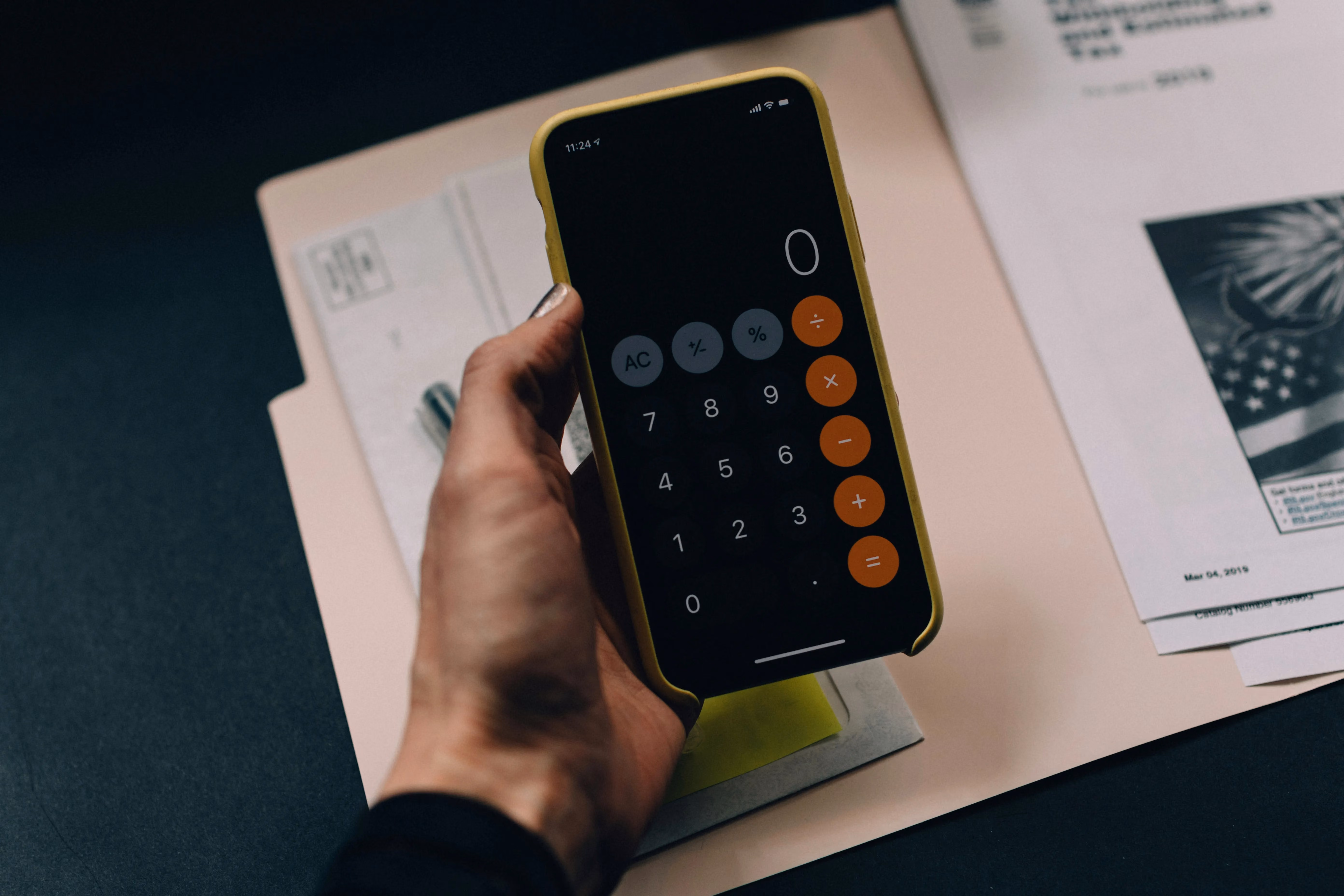 Hand hält ein Smartphone mit geöffneter Taschenrechner-App über einem Ordner und Papieren.