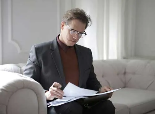 Geschäftsmann mit Brille sitzt auf Sofa und überprüft Papiere in einem Aktenordner.