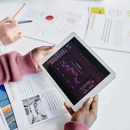 Person hält Tablet mit buntem Diagramm, andere Person zeigt mit Stift auf Papiere mit Grafiken.