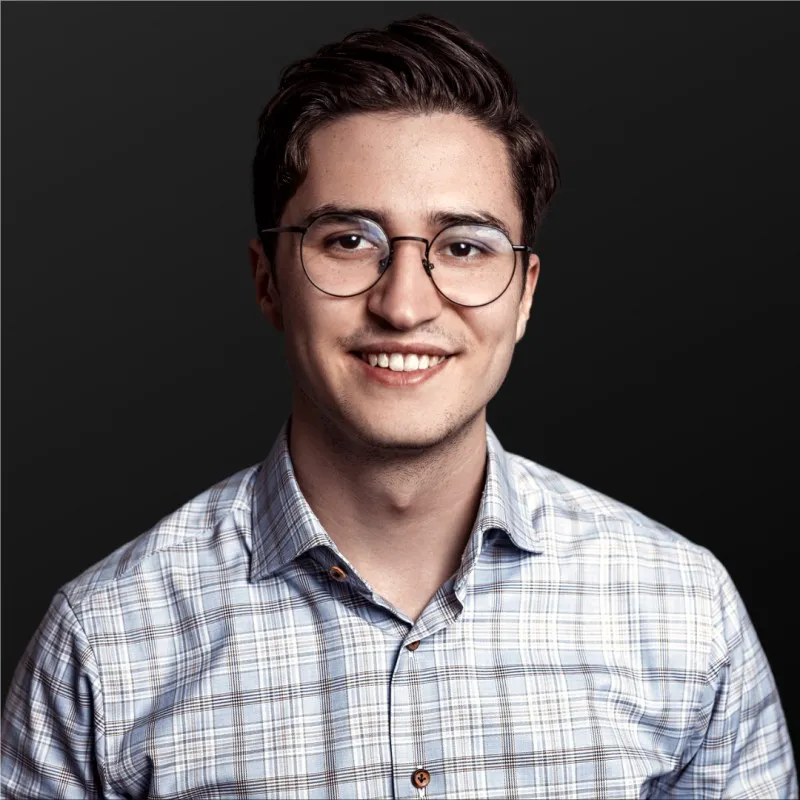 Junger Mann mit Brille und kariertem Hemd vor dunklem Hintergrund lächelt.