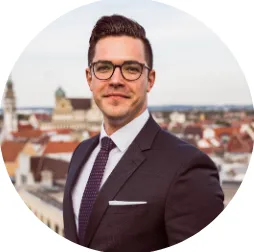 Junger Mann mit Brille in formellem Anzug vor einer Stadtansicht im Hintergrund.