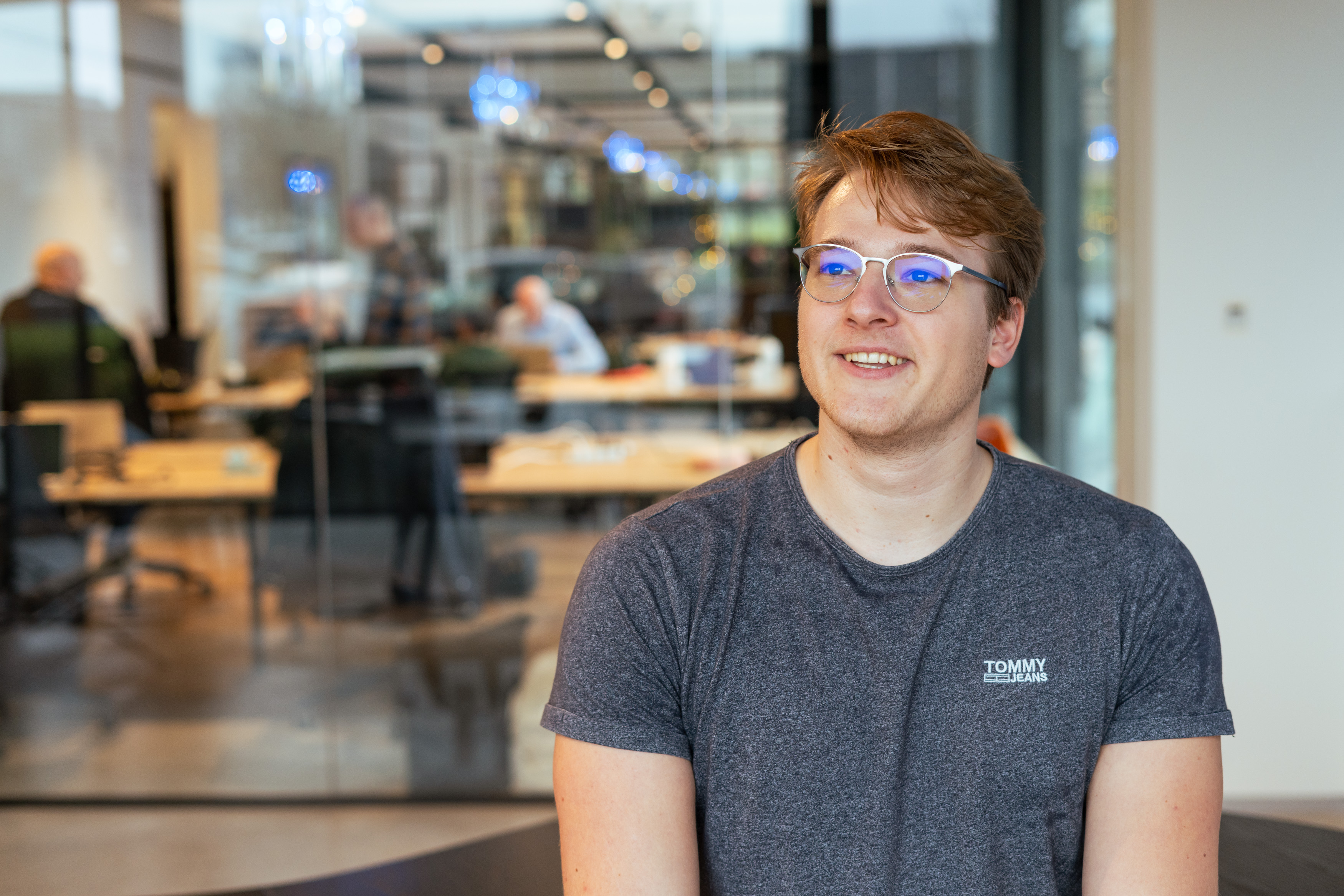 Smiling young man with glasses and a gray Tommy Jeans t-shirt seated indoors with an office space visible in the background.