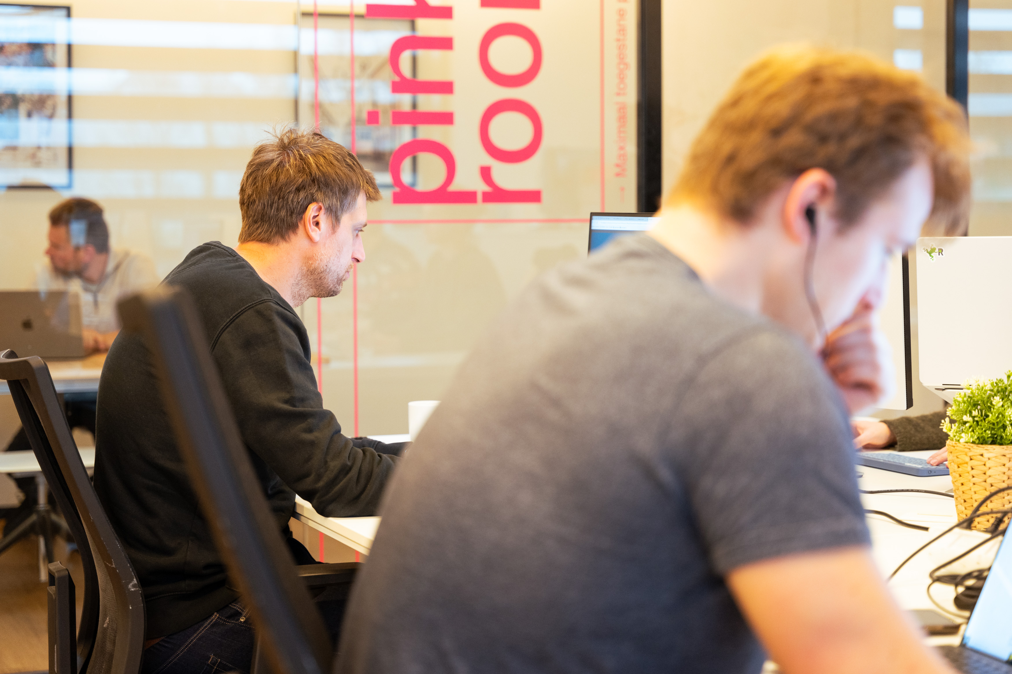 Man wearing orange and black headset sitting at a desk with a laptop in a modern office.