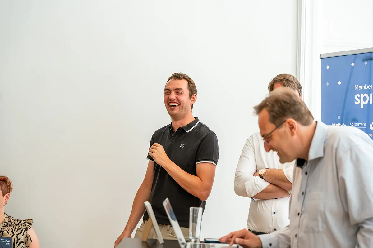 Smiling young man with glasses and a gray Tommy Jeans t-shirt seated indoors with an office space visible in the background.