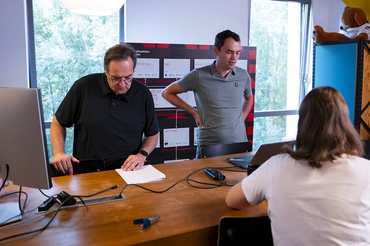 Smiling young man with glasses and a gray Tommy Jeans t-shirt seated indoors with an office space visible in the background.
