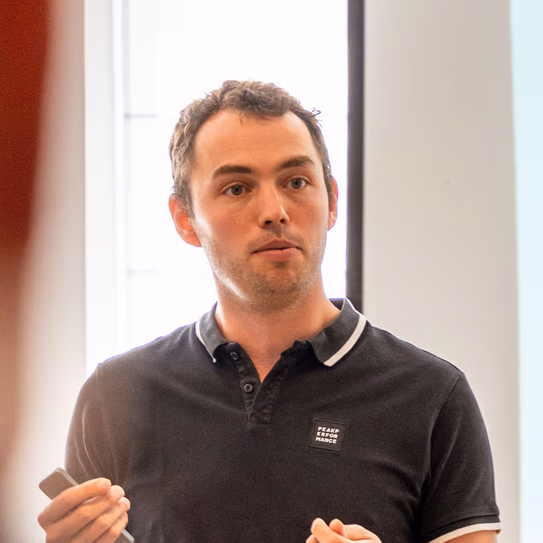 Man in a black polo shirt holding a remote, speaking indoors with a window in the background.