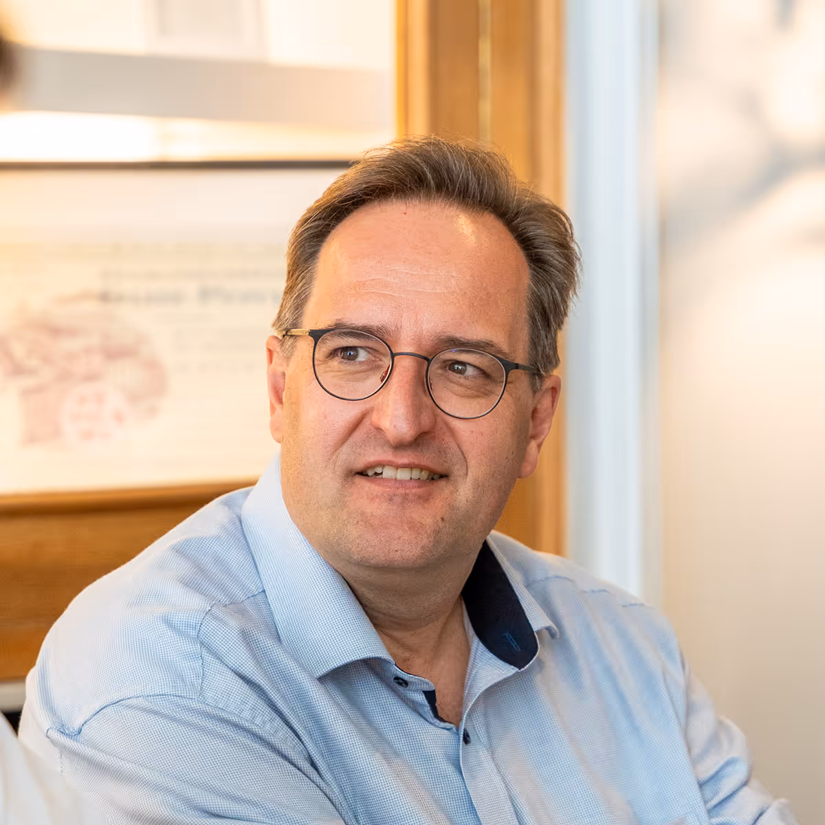 Middle-aged man with glasses wearing a light blue shirt looking to the side in a warmly lit room.