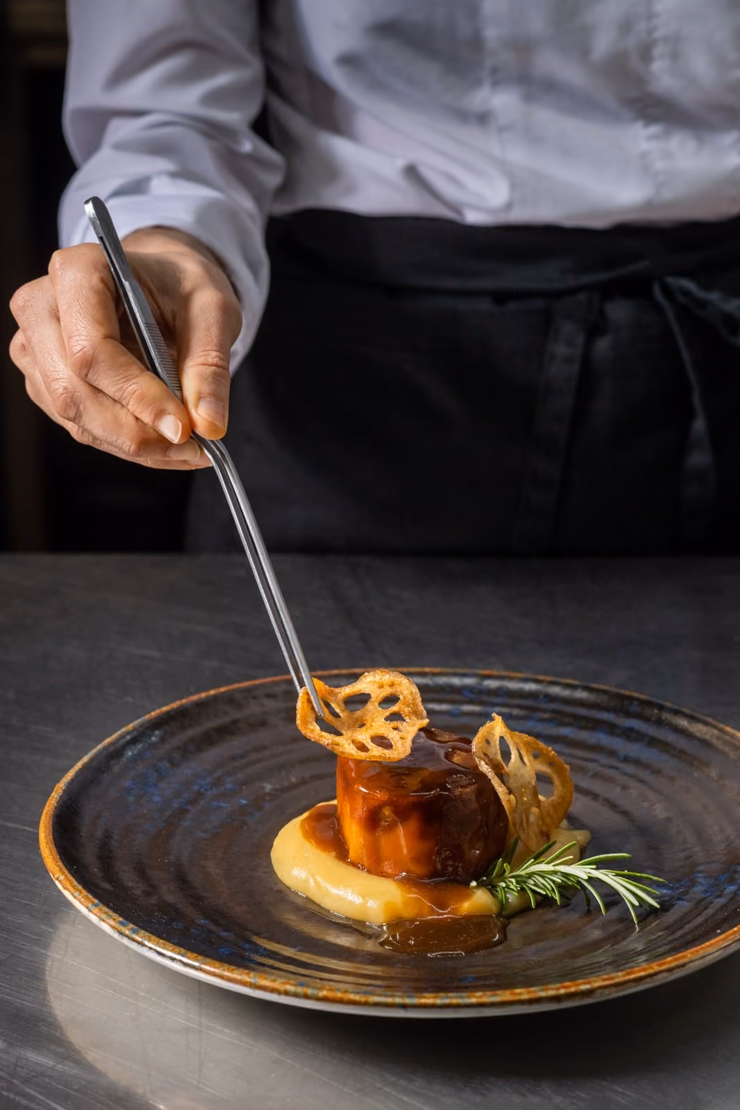 Manos de chef colocando una guarnición crujiente en un plato gourmet con puré y salsa.