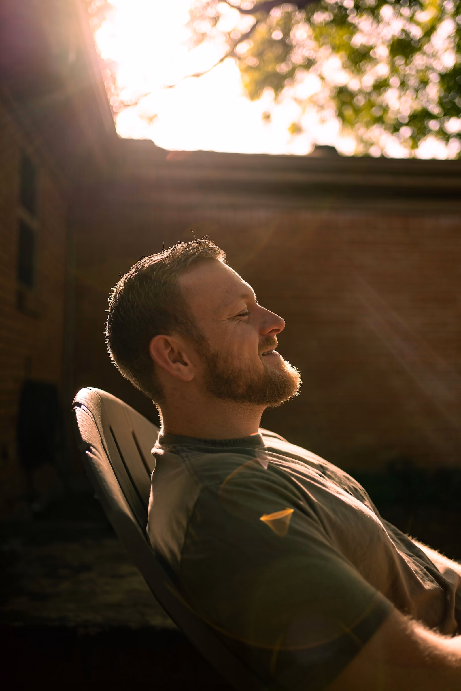 Hombre relajado sentado en silla al sol con ojos cerrados y sonrisa suave.