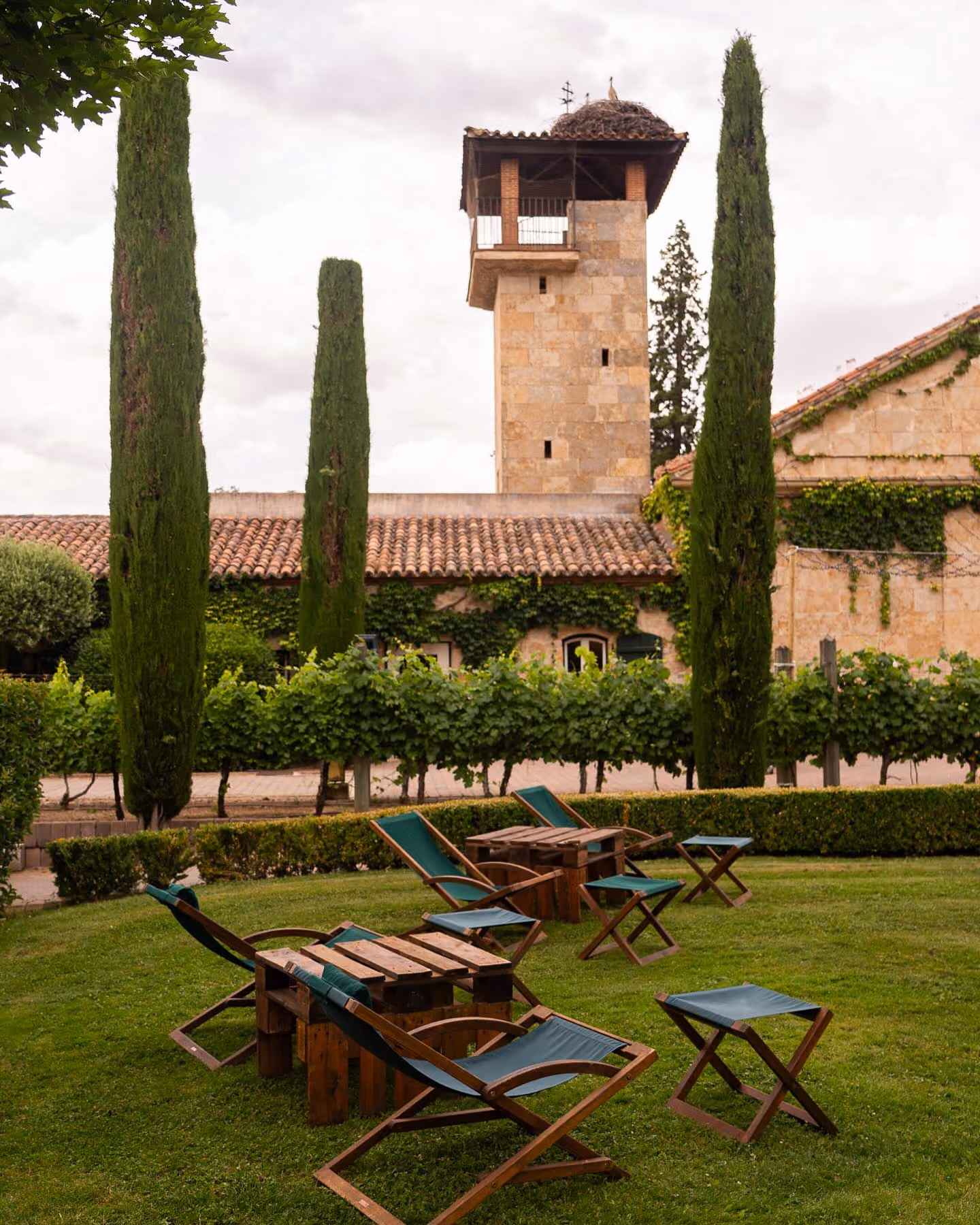 Jardín verde con sillas reclinables y mesa de madera frente a edificio de piedra con torre.
