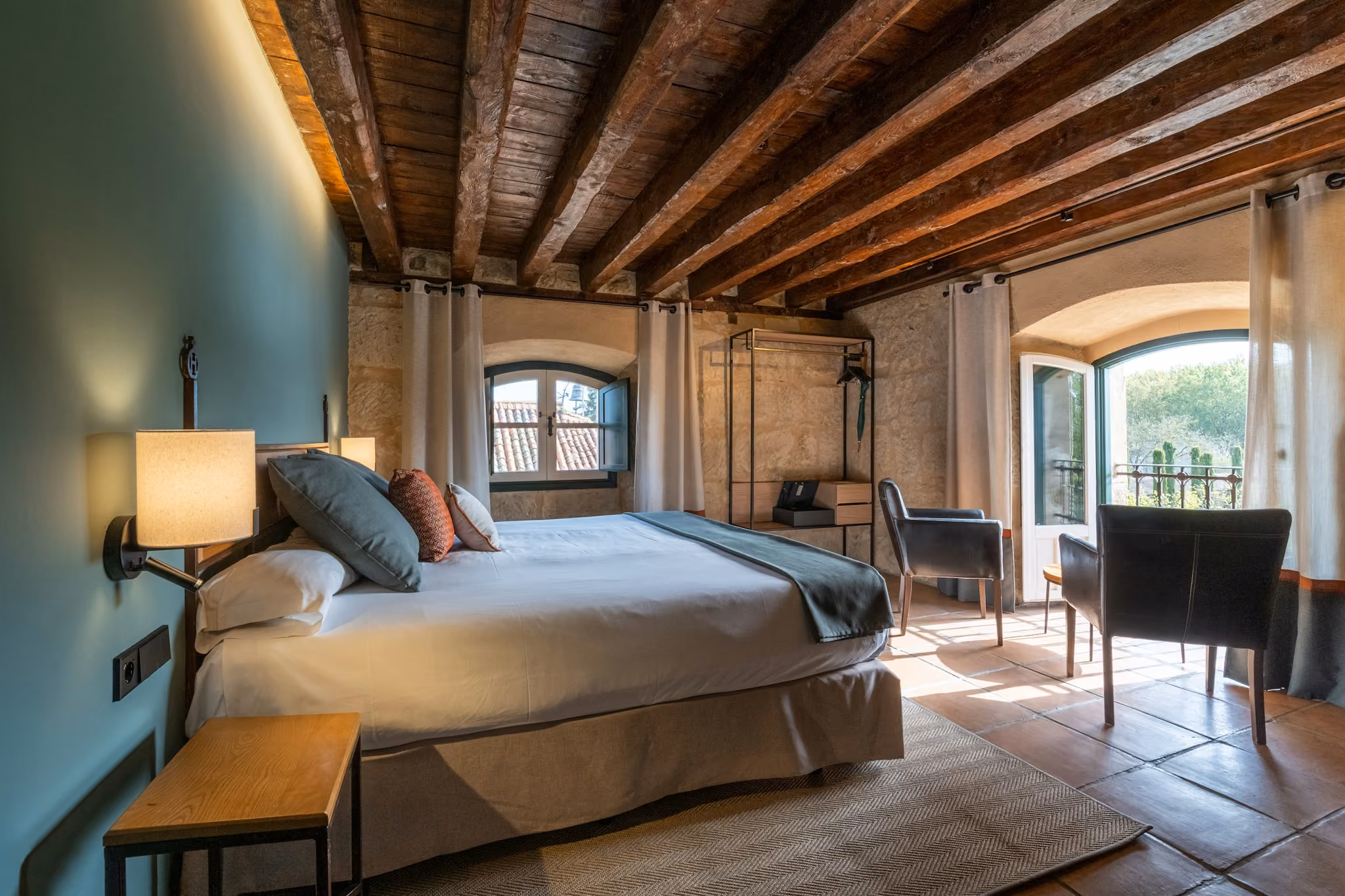 Habitación con cama grande, paredes de piedra, techo de vigas de madera y ventanales con vista al exterior.