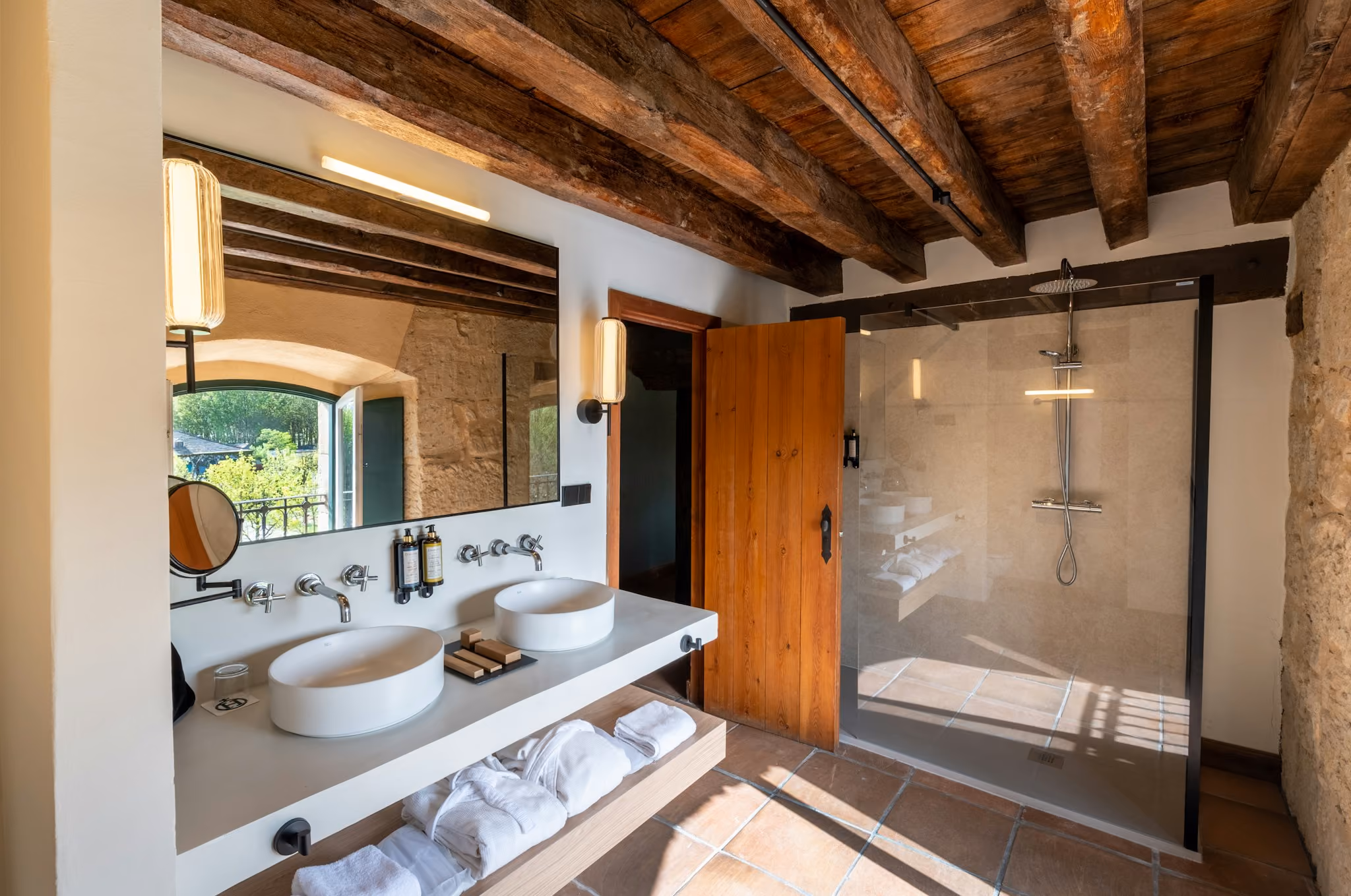 Baño con lavabos dobles, ducha con mampara de vidrio, techo con vigas de madera y ventana abierta.