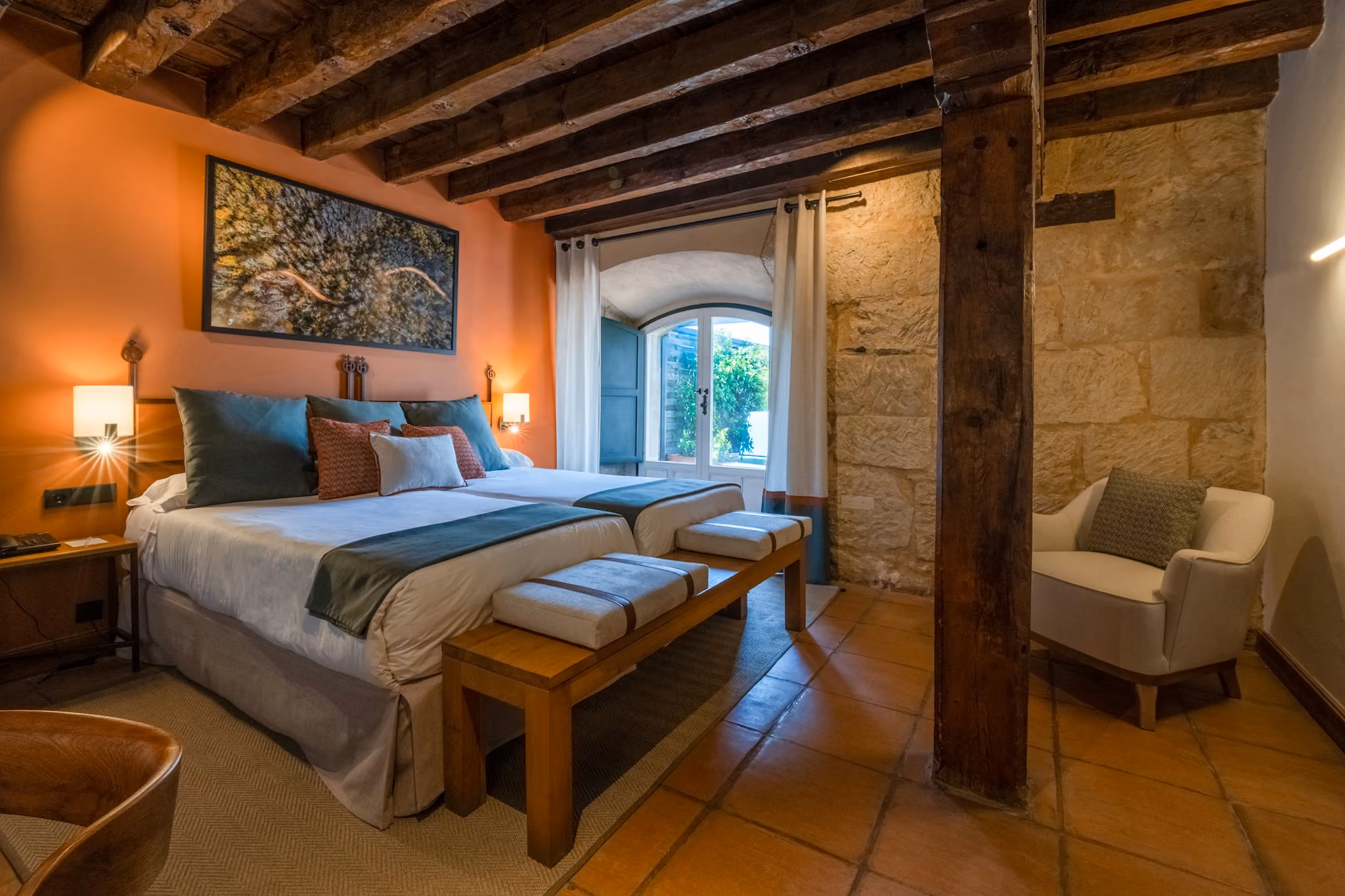 Habitación con dos camas, vigas de madera, pared de piedra, ventana con cortinas y silla blanca.