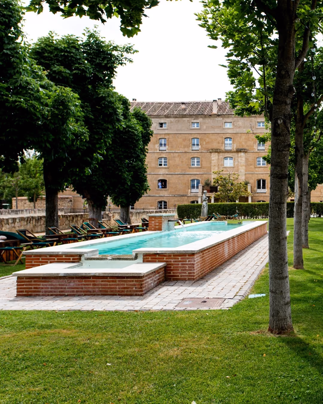 Piscina alargada con borde de ladrillos y tumbonas junto a árboles y edificio antiguo al fondo.