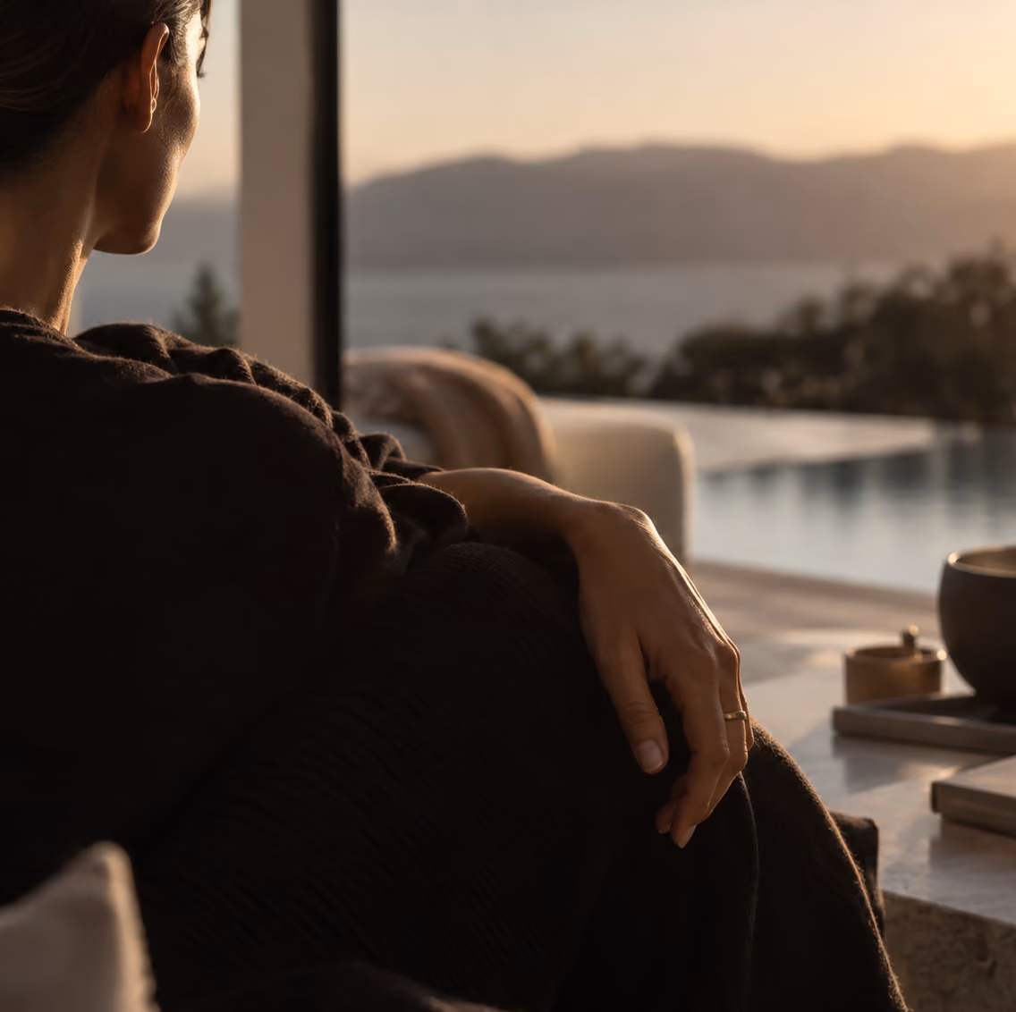 Mujer vista de perfil sentada en un interior, mirando por una ventana hacia un paisaje con montañas y un cuerpo de agua al atardecer.