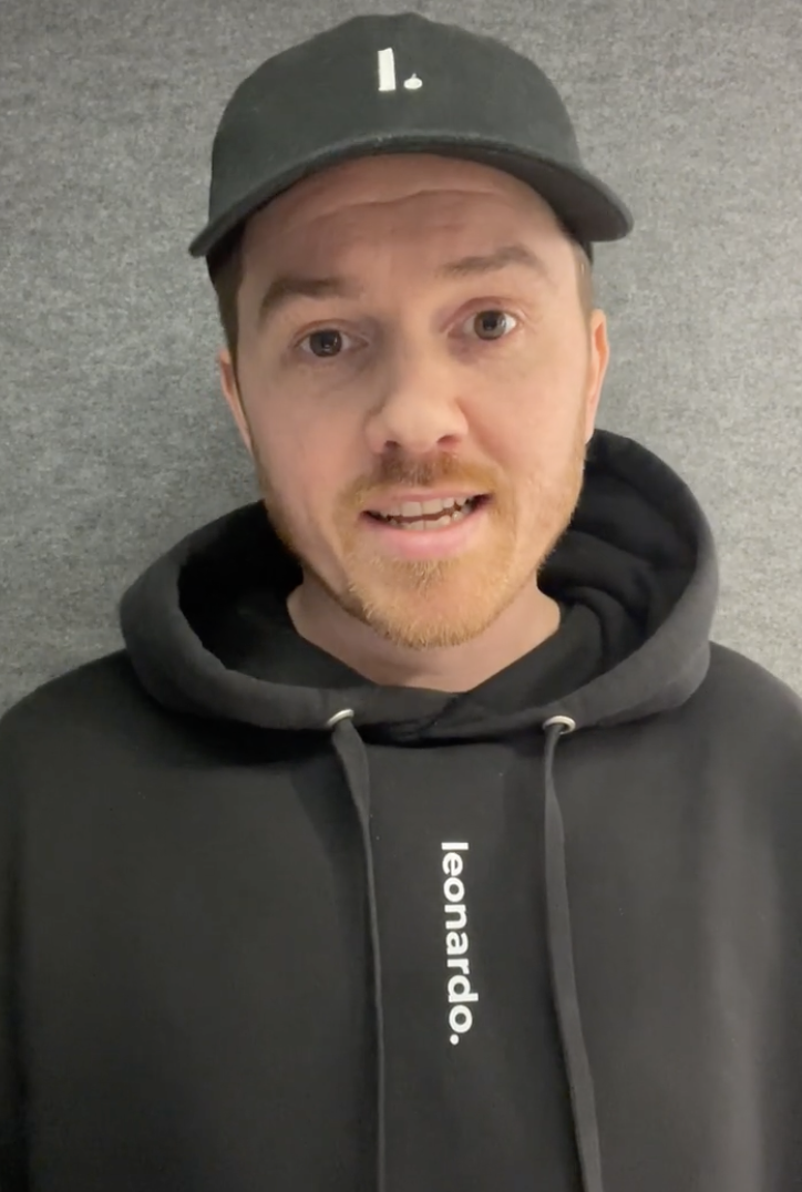Man with light facial hair wearing a black cap and black hoodie with 'leonardo.' printed on it, standing against a gray background.