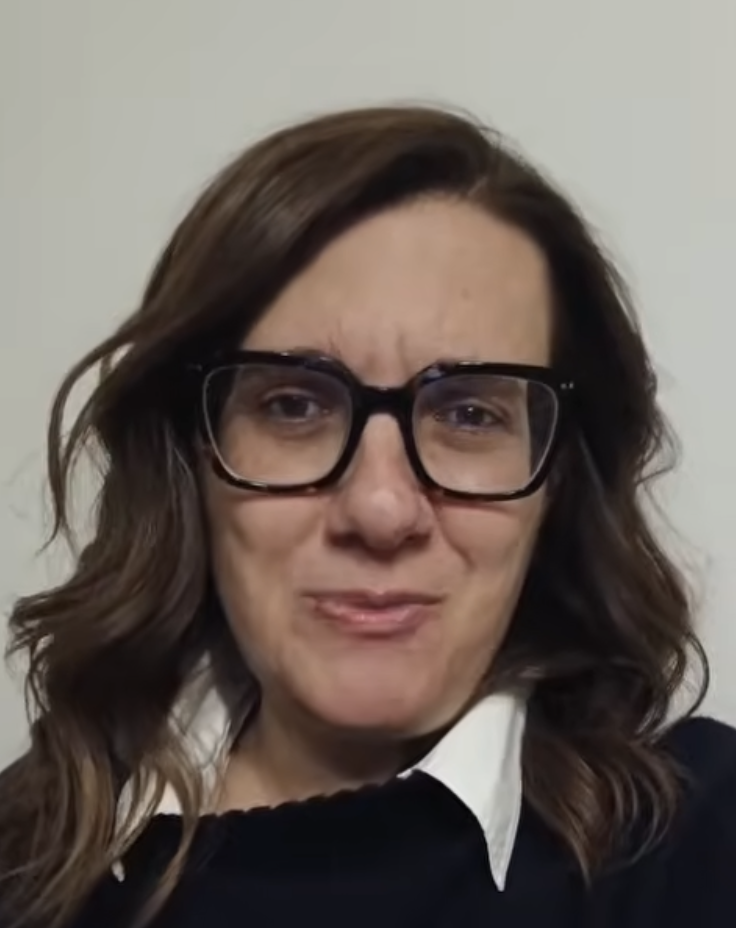 Woman with wavy brown hair wearing large black glasses, a white collared shirt, and a dark sweater, looking at the camera with a subtle smile.
