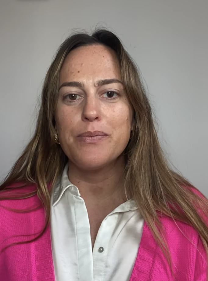 Woman with long brown hair wearing a white shirt and bright pink cardigan against a plain gray background.