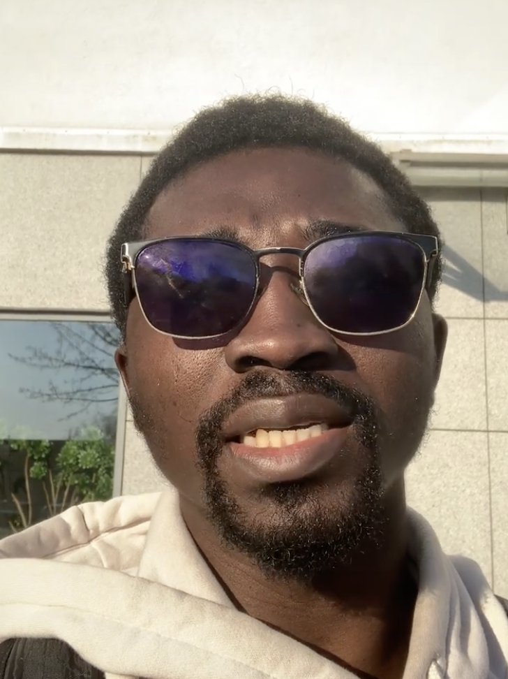 Close-up of a man with short curly hair wearing sunglasses and a light-colored hoodie, standing outdoors in sunlight.