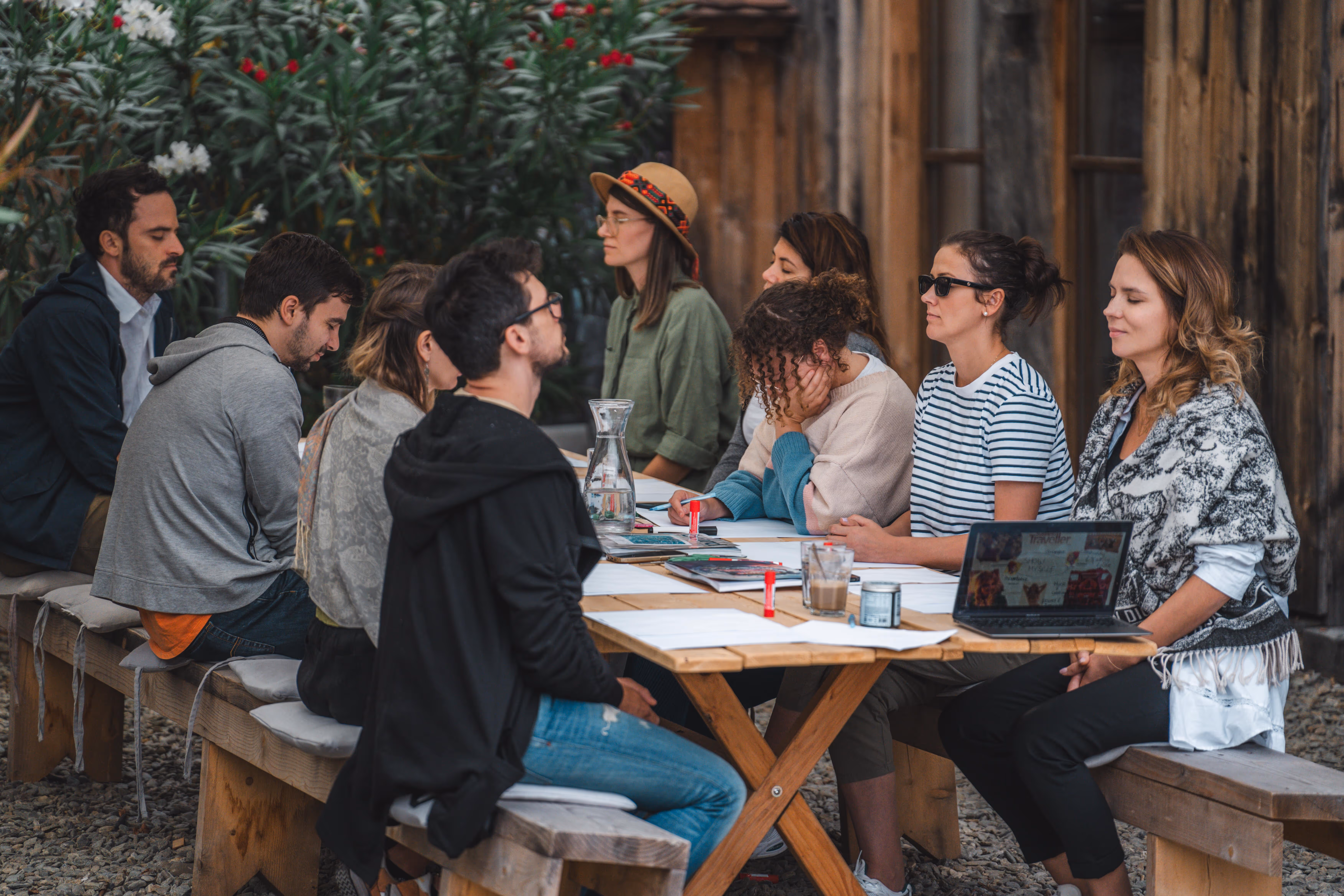 Group of eight people sitting around a wooden table outdoors, appearing to meditate or relax with eyes closed.