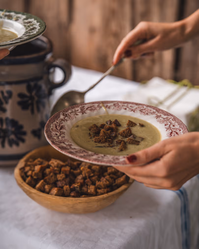 Hands holding a plate of creamy soup topped with croutons, with a wooden bowl of extra croutons and a ceramic pitcher in the background.