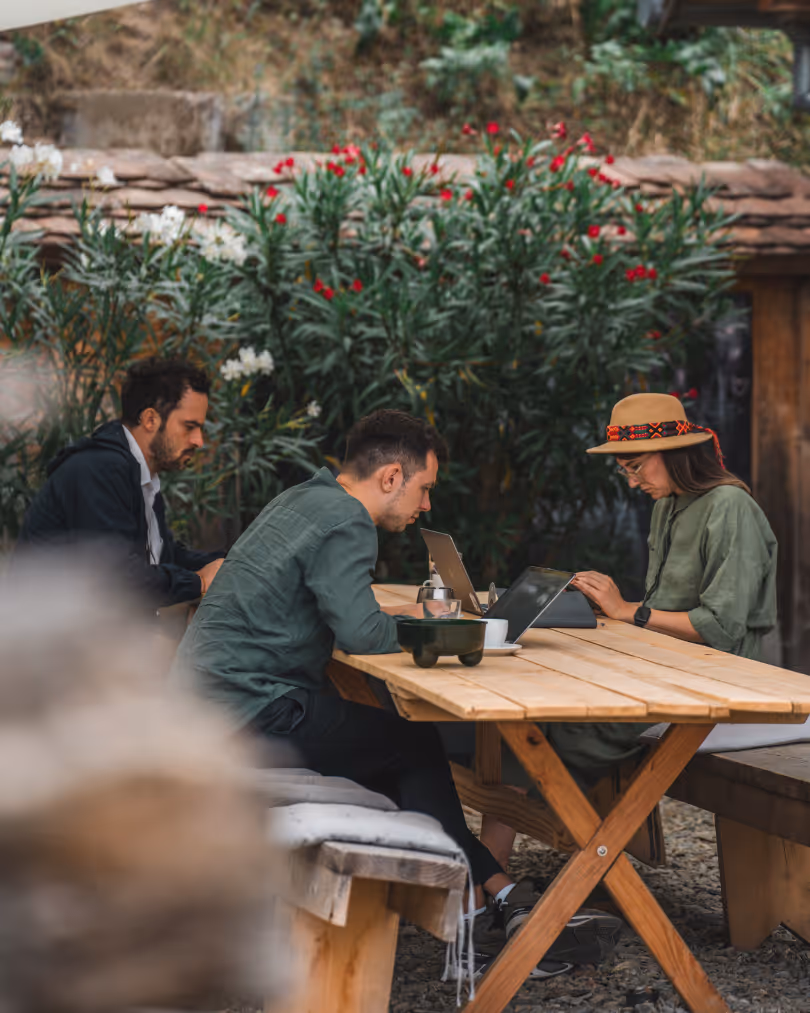 Three people sitting outdoors at a wooden picnic table working on laptops surrounded by greenery with red and white flowers.
