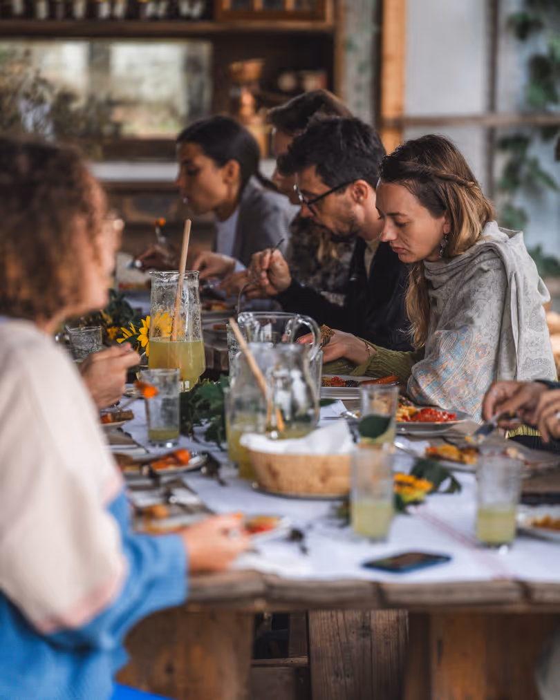 Group of people seated at a rustic wooden table enjoying a meal together, with pitchers of lemonade and plates of food.