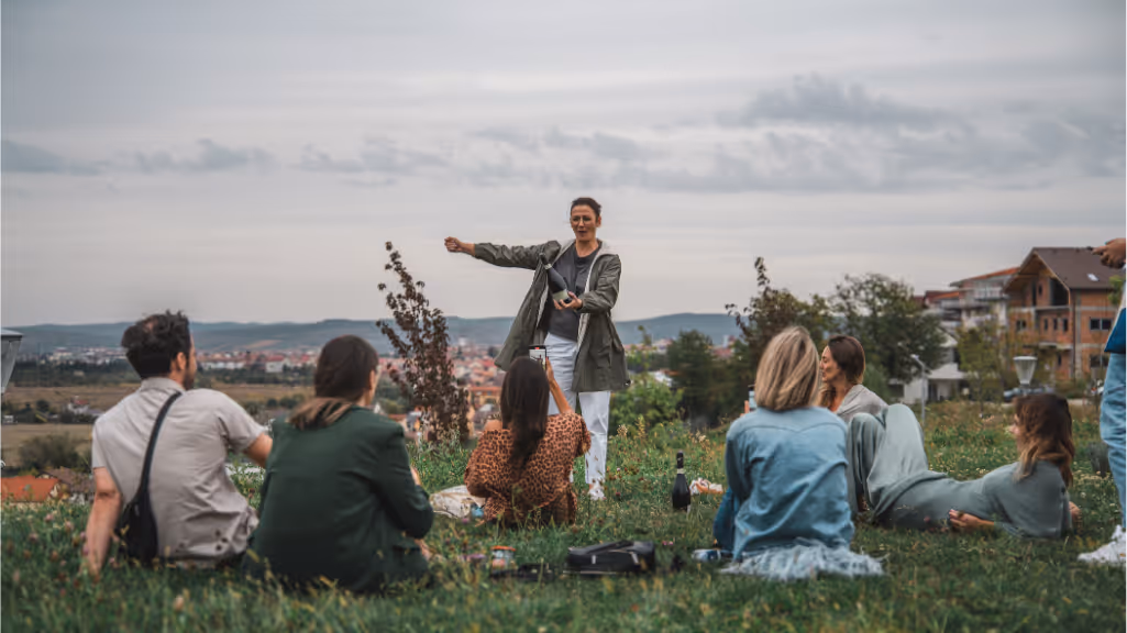 Group of people sitting on grass outdoors with one person standing and opening a bottle, under a cloudy sky.