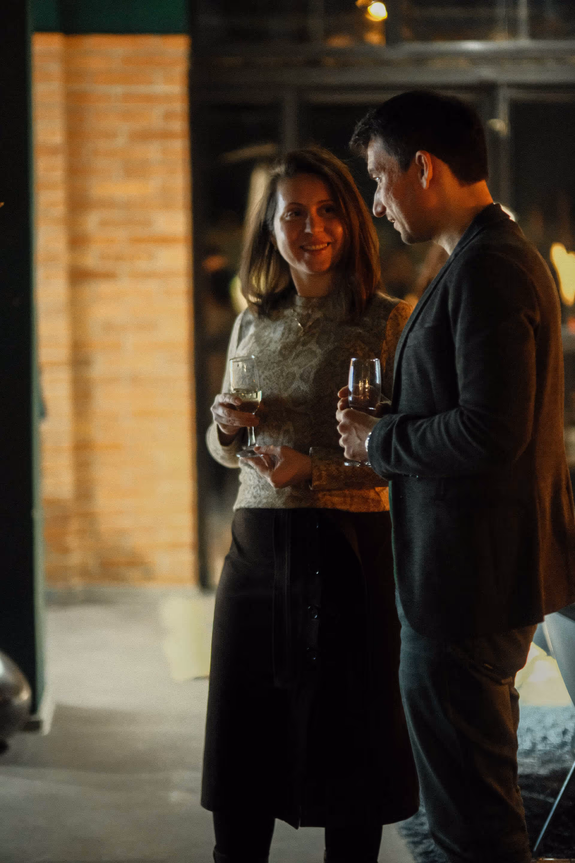 A man and a woman holding wine glasses while having a conversation in a dimly lit indoor setting.